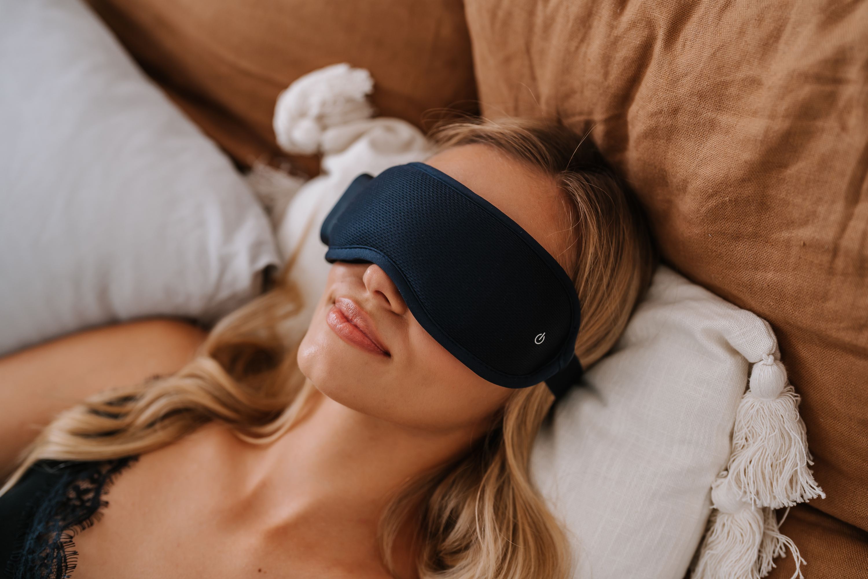 Frau mit einer marineblauen Schlafmaske, die auf einem Bett mit Kissen liegt.