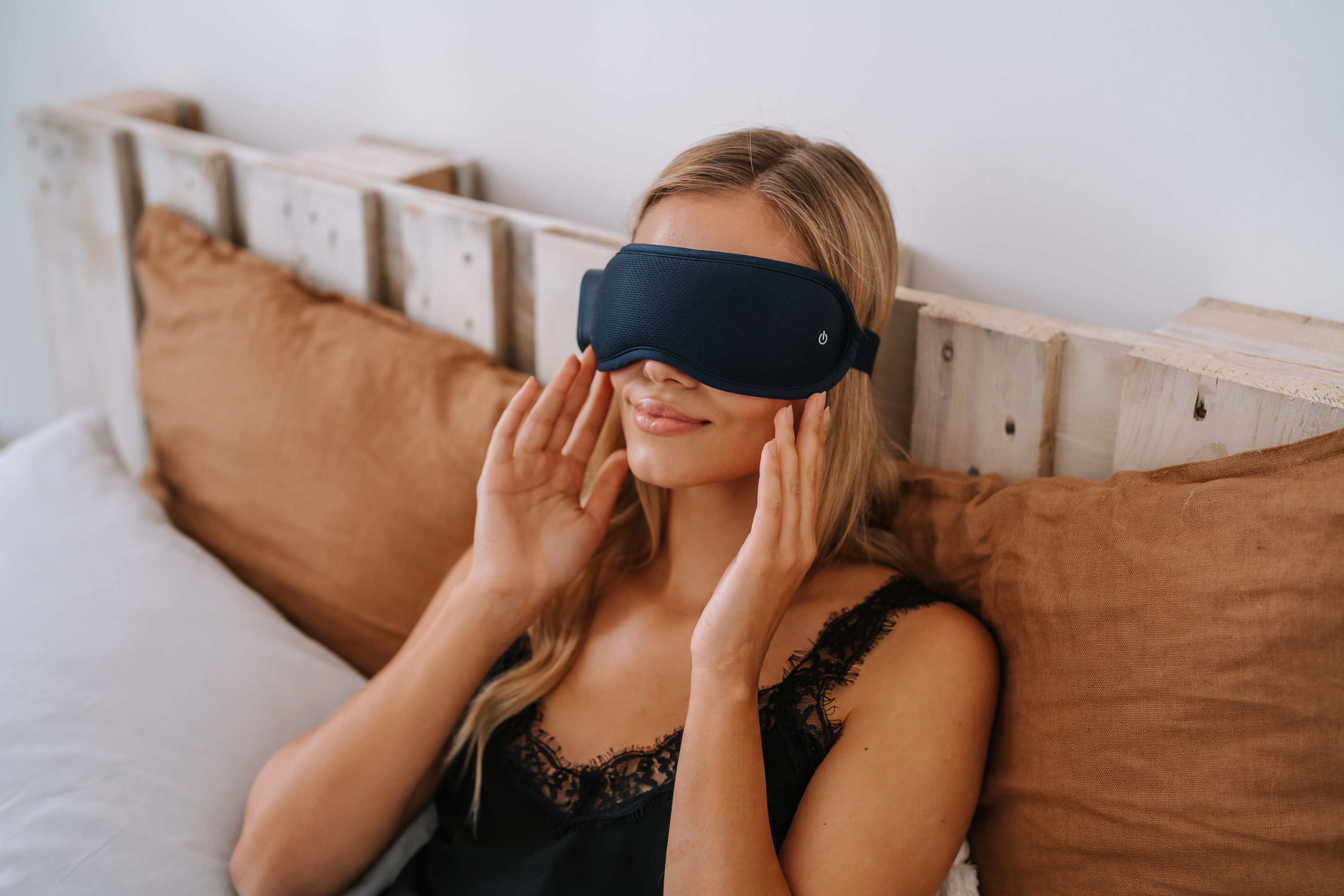Frau mit schwarzer Schlafmaske, Hände auf der Maske, sitzt auf Bett mit Kissen.