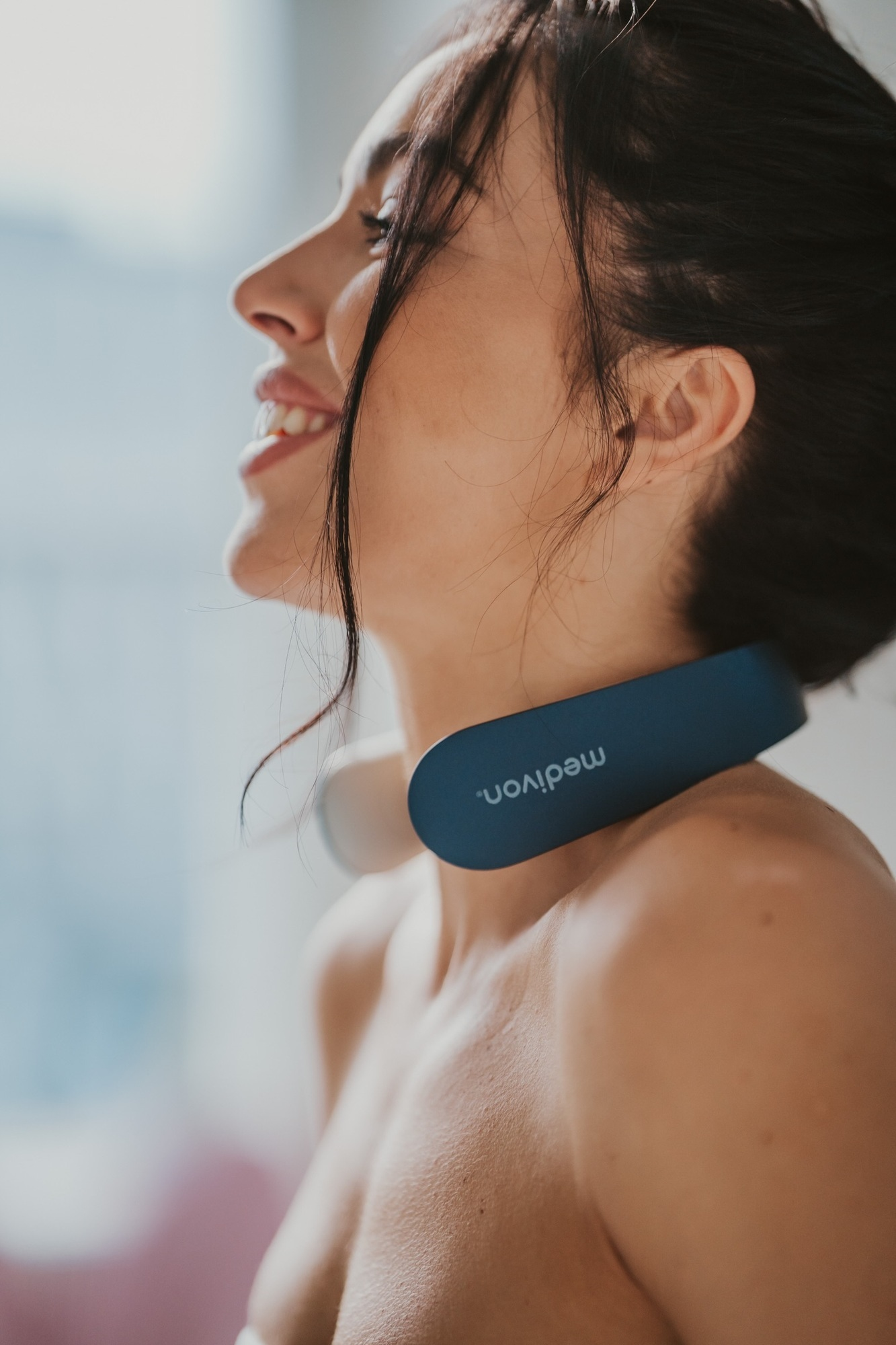 Eine Frau mit einem blau-weißen Nackenmassagegerät, lächelnd, in unscharfer Umgebung.