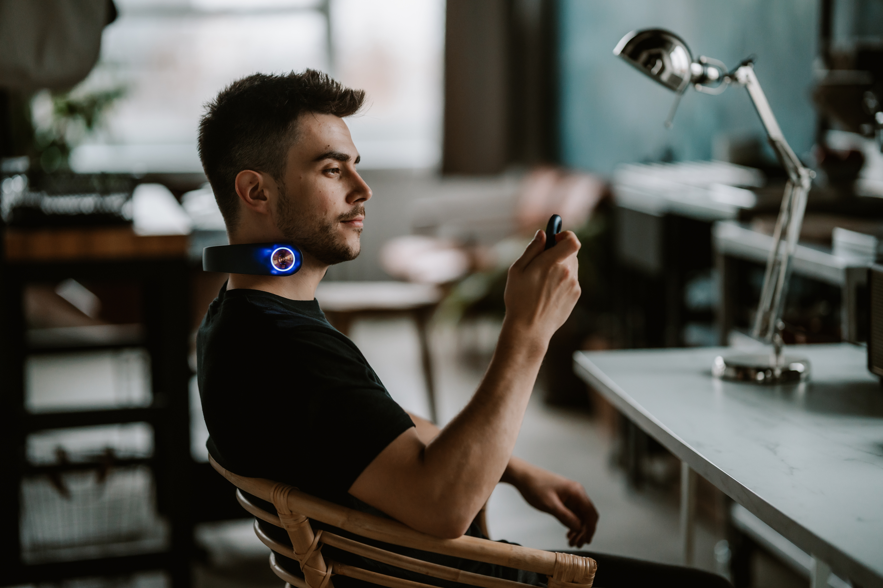 Mann mit Nackenmassagegerät, hält Fernbedienung. Innenansicht mit Schreibtischlampe.