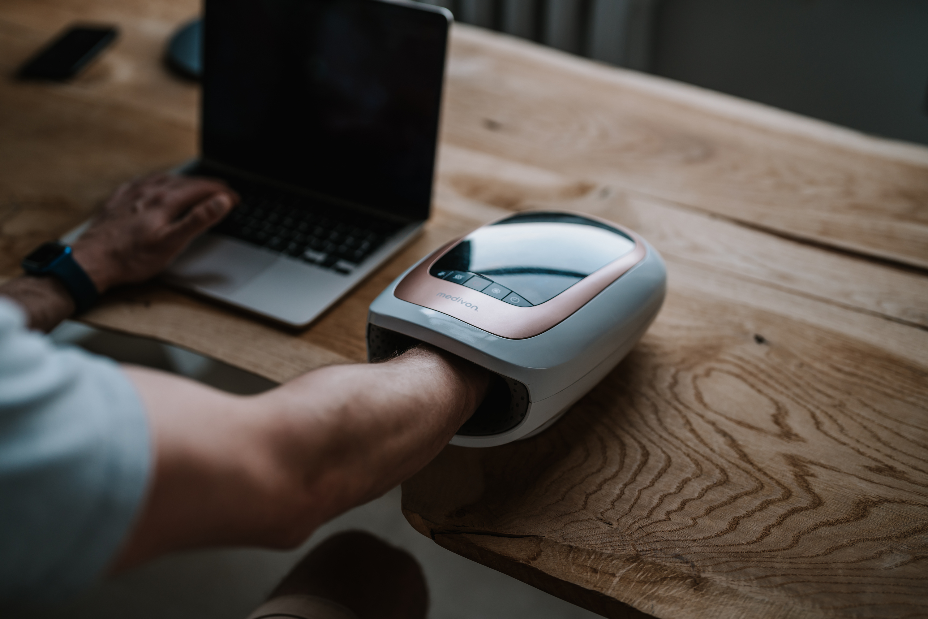 Person benutzt Handmassagegerät neben Laptop auf Holztisch.