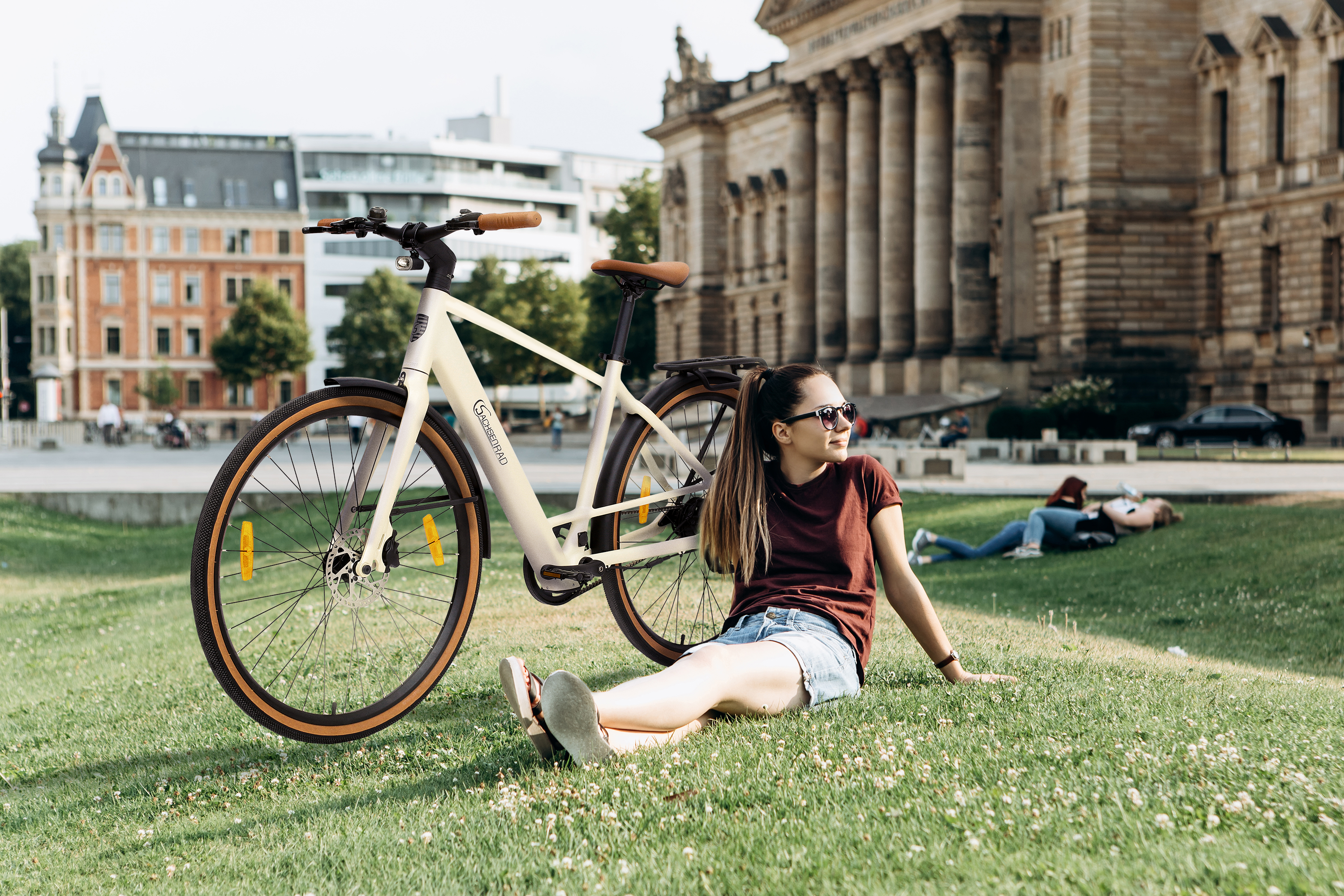 Eine Frau sitzt neben einem cremefarbenen Fahrrad. Ein Gebäude befindet sich im Hintergrund. Andere Leute sind auf dem Rasen.