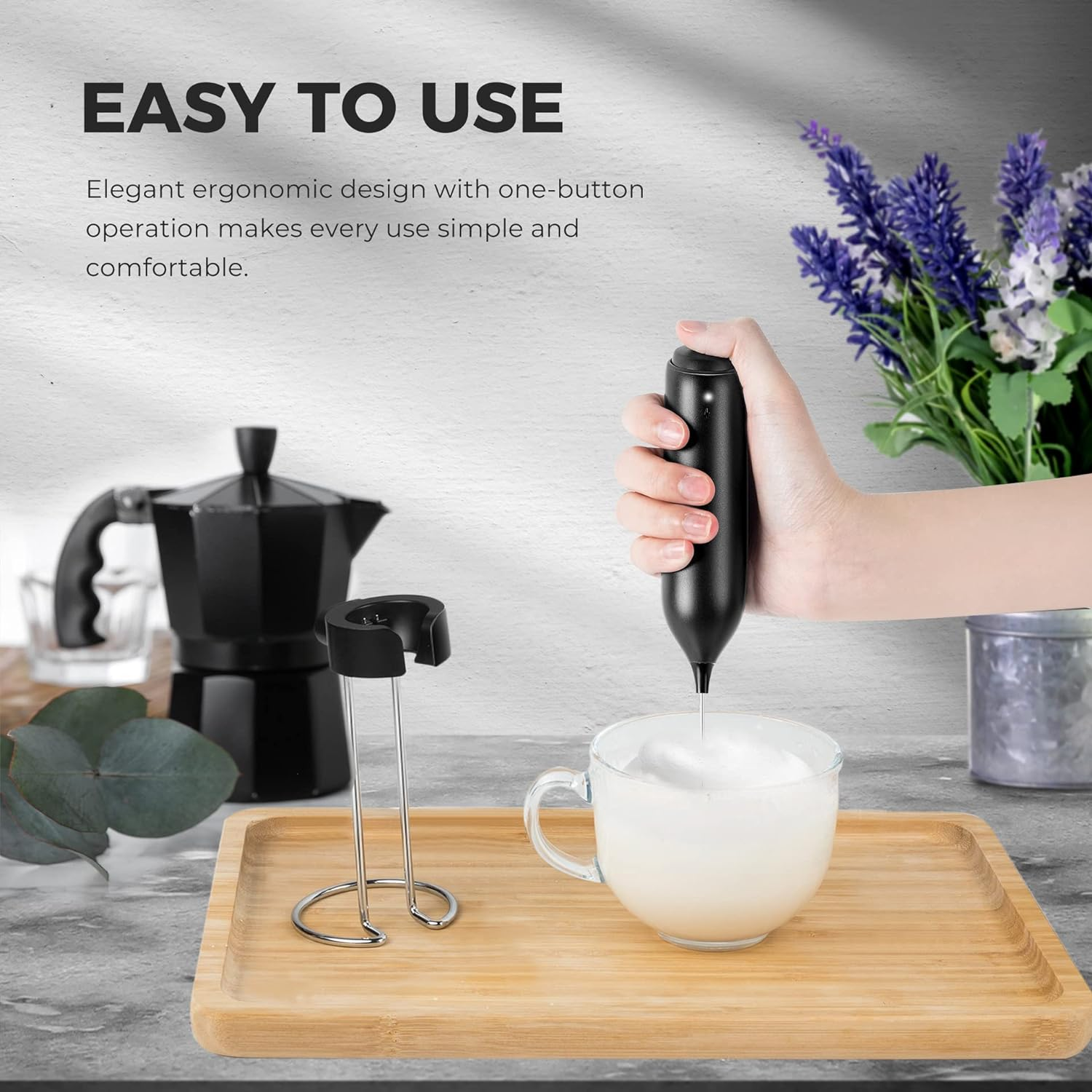 Eine Hand hält einen schwarzen Milchaufschäumer, der Milch in einem Glas aufschäumt, mit Tablett und Kaffeekanne.