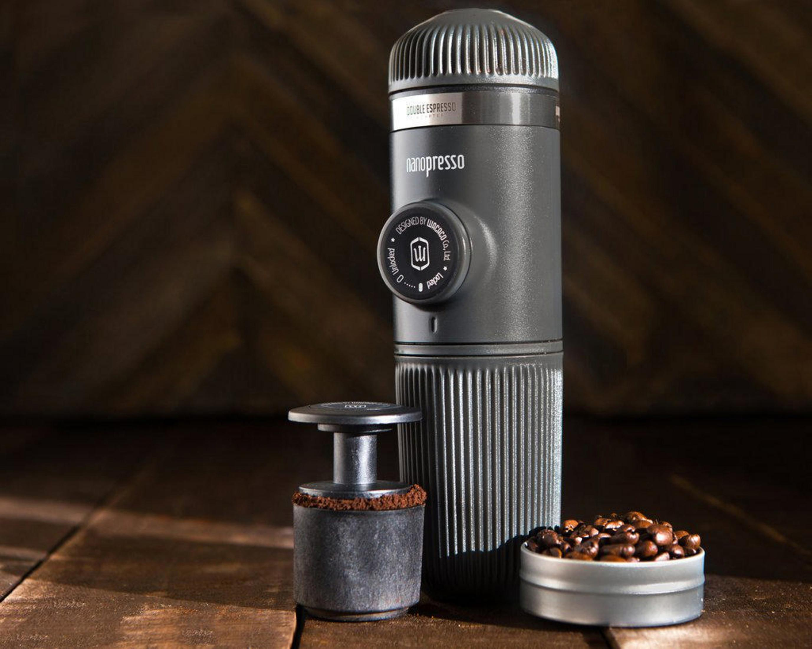 Cafetera espresso portátil con granos de café y café molido, sobre una superficie de madera.