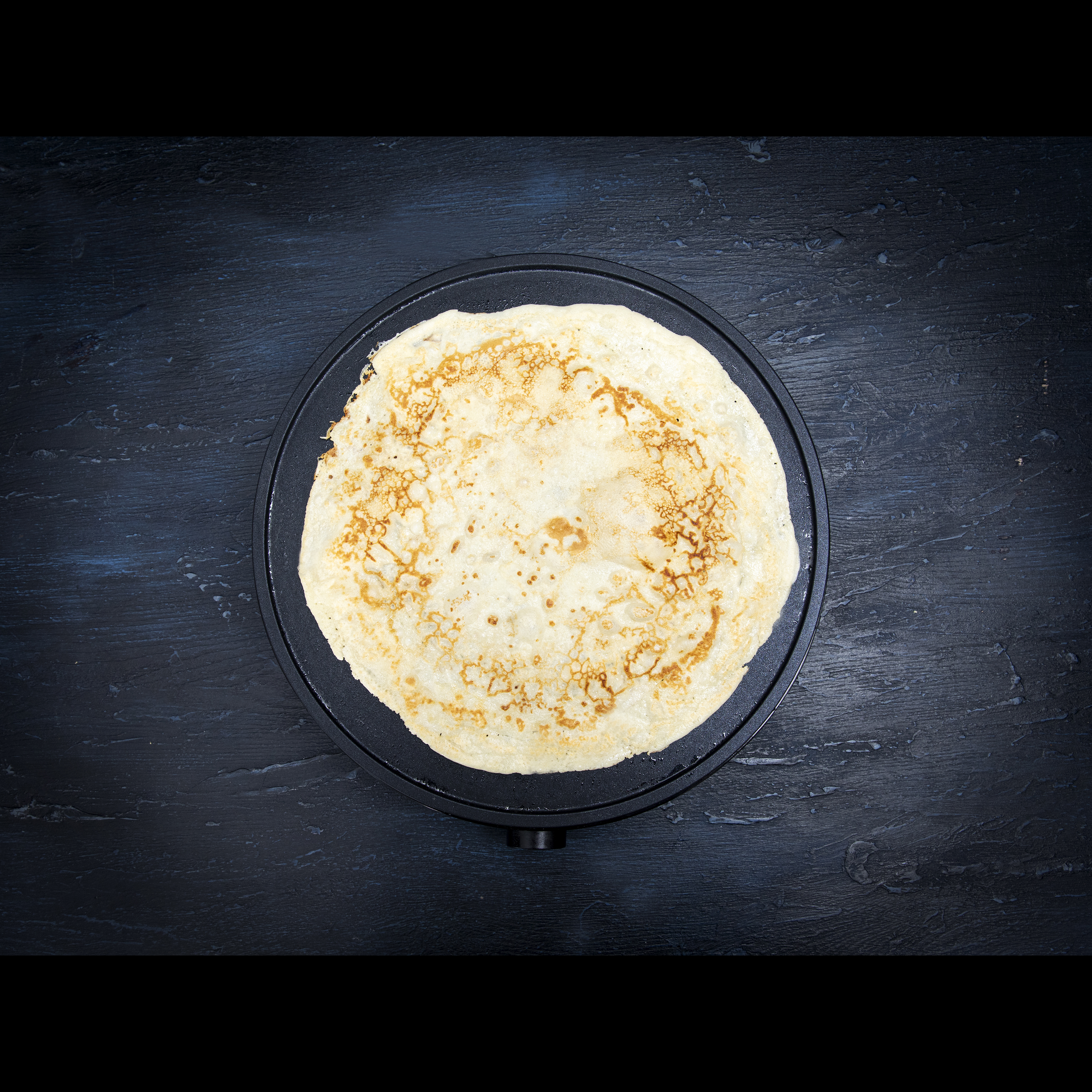 Naleśnik na czarnej płycie grzejnej. Naleśnik jest okrągły i złocistobrązowy.