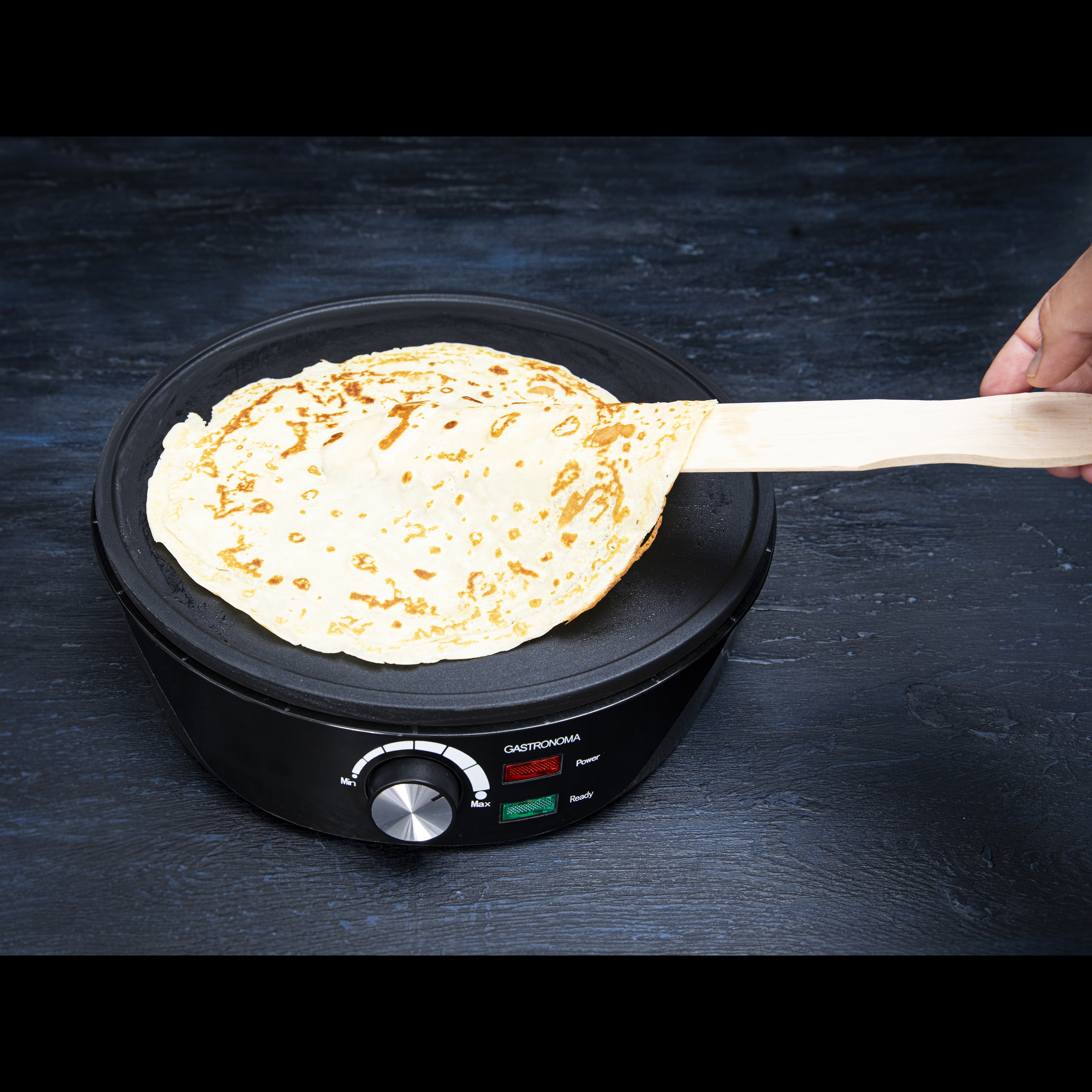Naleśnik na czarnej płycie grzejnej, trzymany drewnianą szpatułką. Czarna powierzchnia. Dłoń w kadrze.