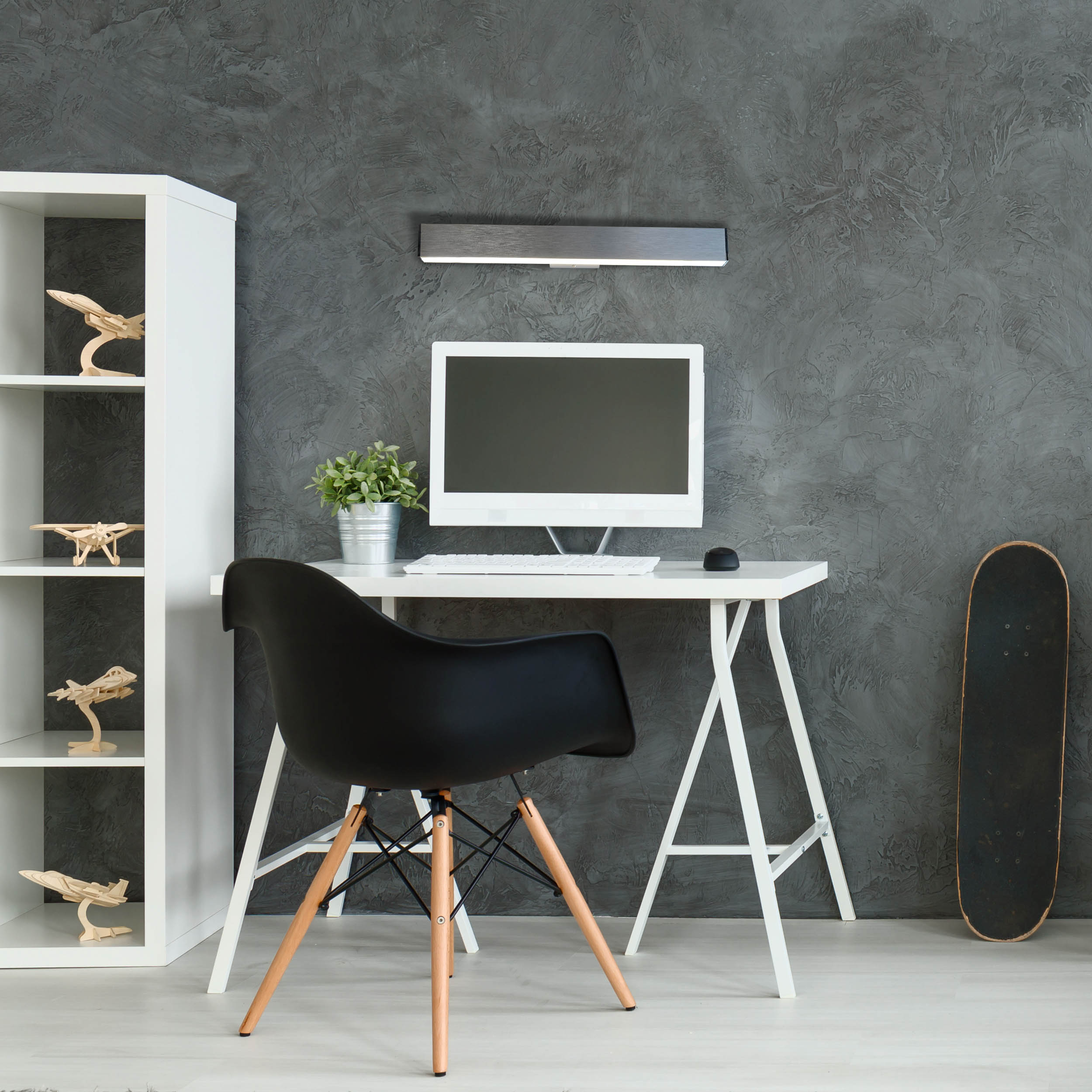 Arbeitsplatz mit weißem Schreibtisch, schwarzem Stuhl, Monitor und Skateboard. Graue Wand.