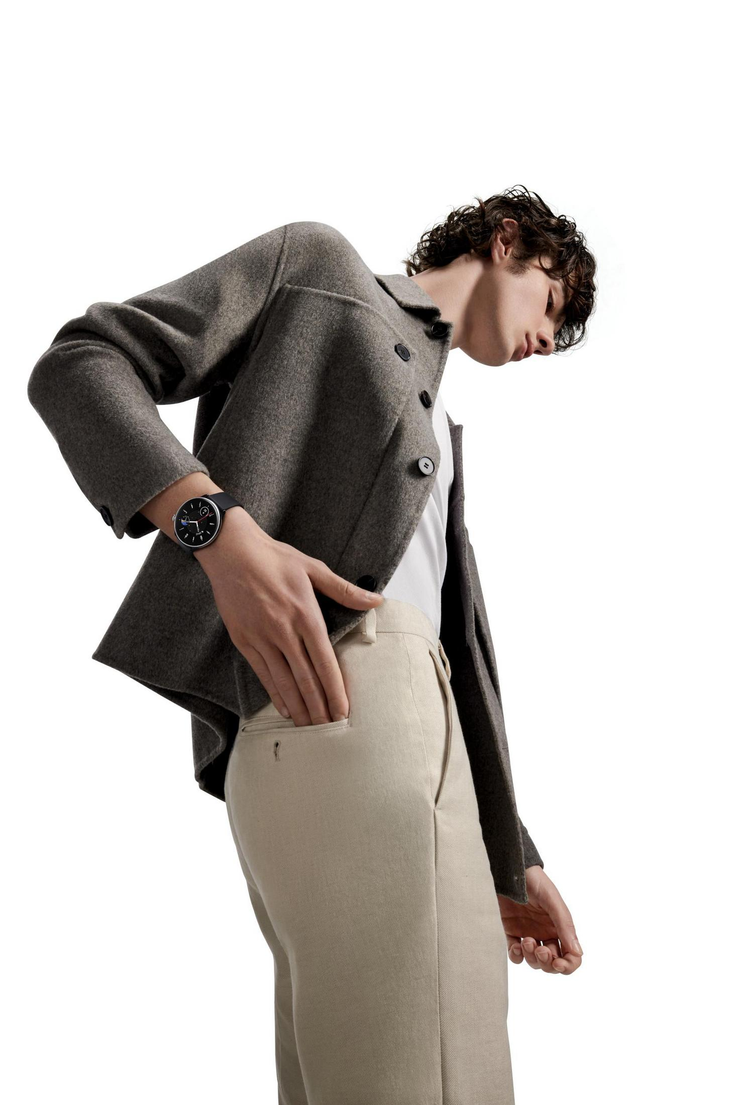 Mężczyzna w kurtce i spodniach, patrzący w dół. Ma zegarek i rękę w kieszeni.