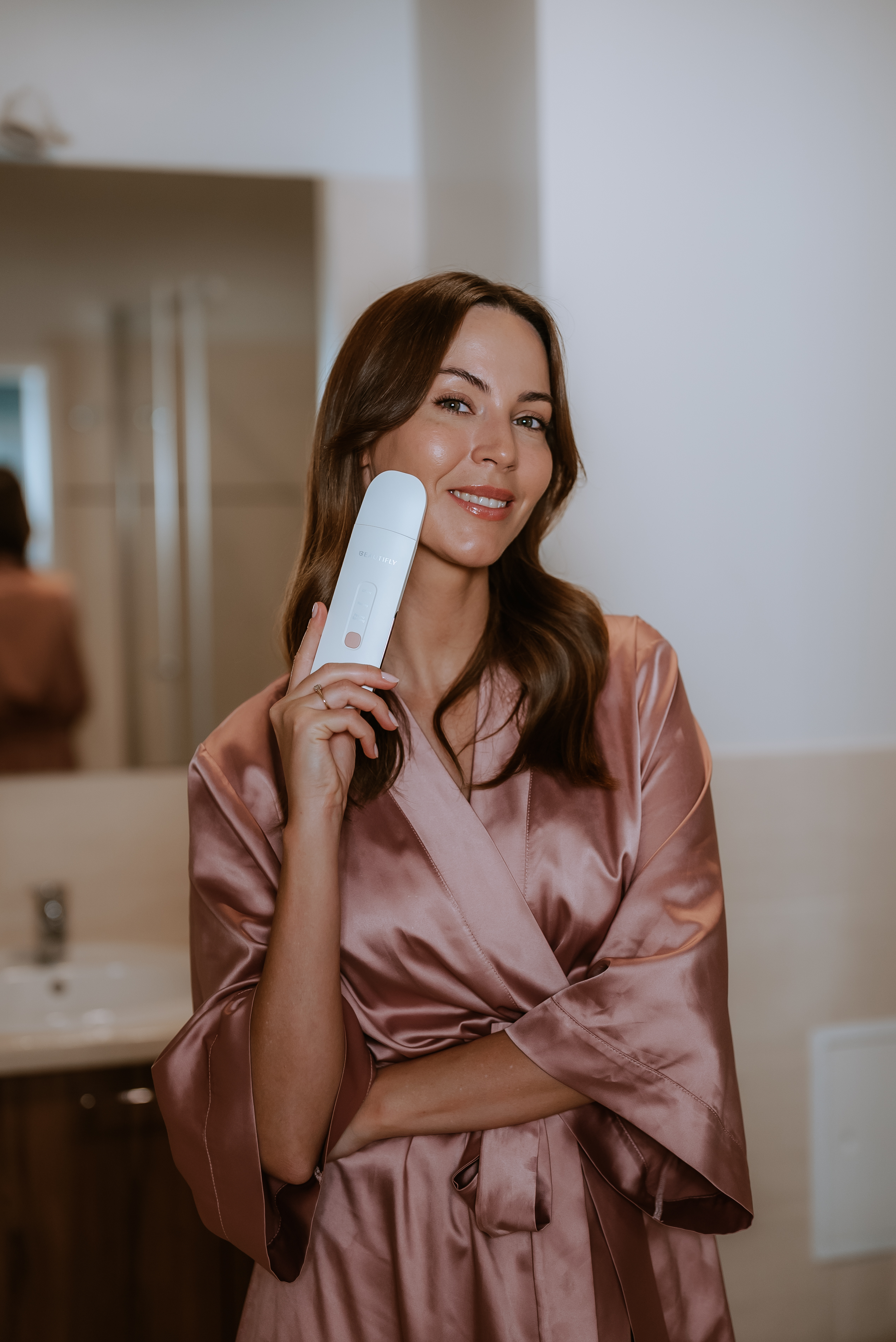 Mujer sosteniendo un dispositivo blanco para el cuidado de la piel en un baño, sonriendo a la cámara.