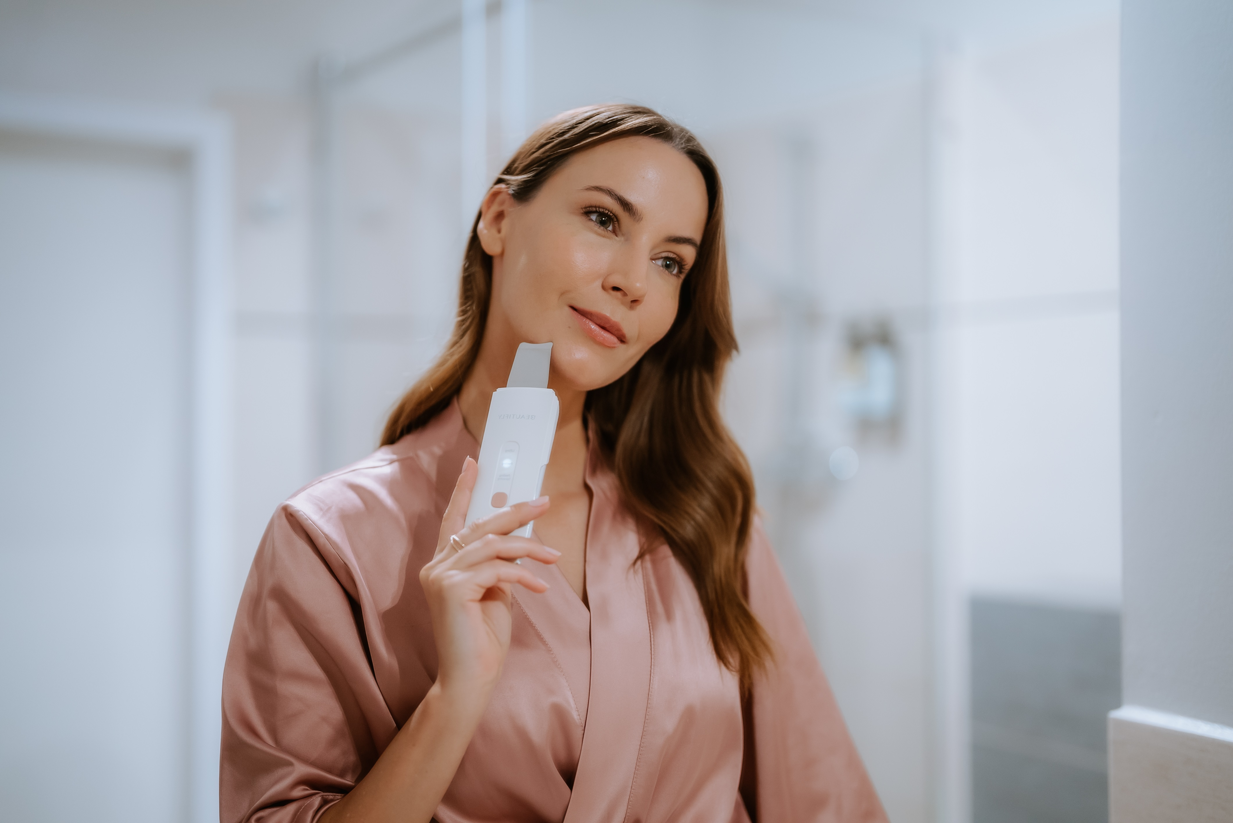 Mujer sosteniendo una herramienta de limpieza facial. Está en un baño, mirándose en un espejo.