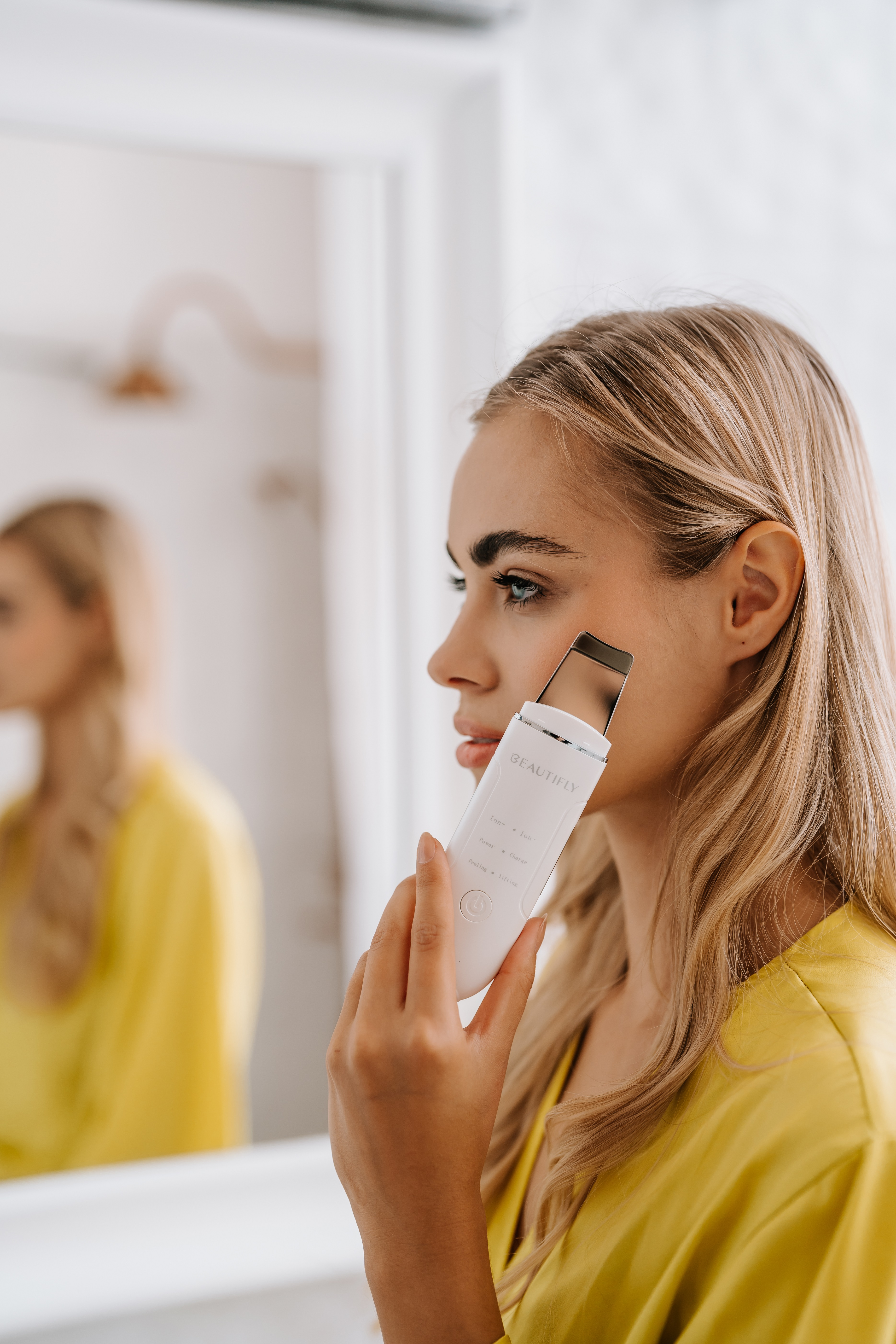 Kobieta w żółtym trzymająca biały skrobak do twarzy przed lustrem. Widoczny odbity obraz.