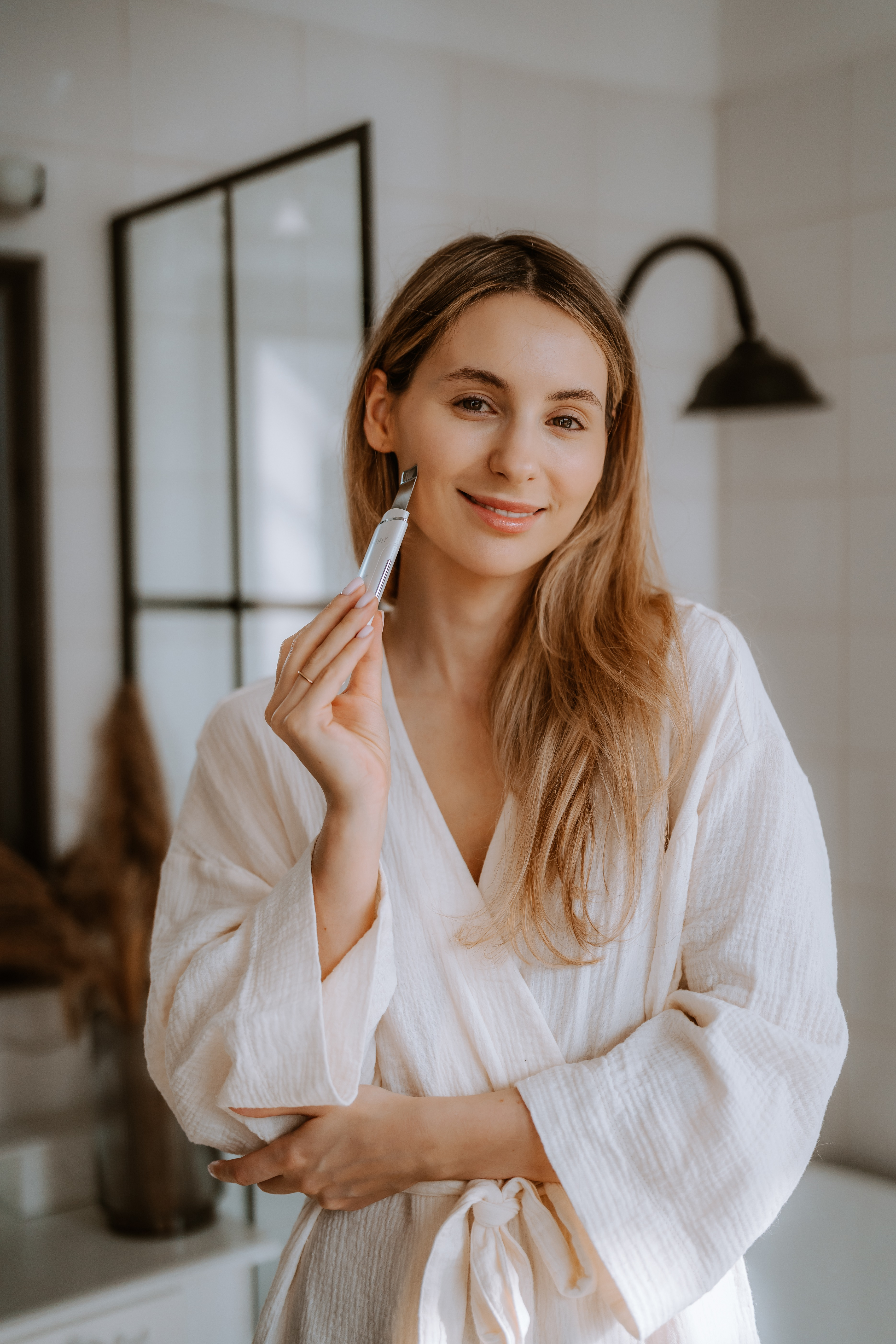 Kobieta w białym szlafroku trzymająca narzędzie do pielęgnacji skóry. Uśmiechnięta, jasne tło.