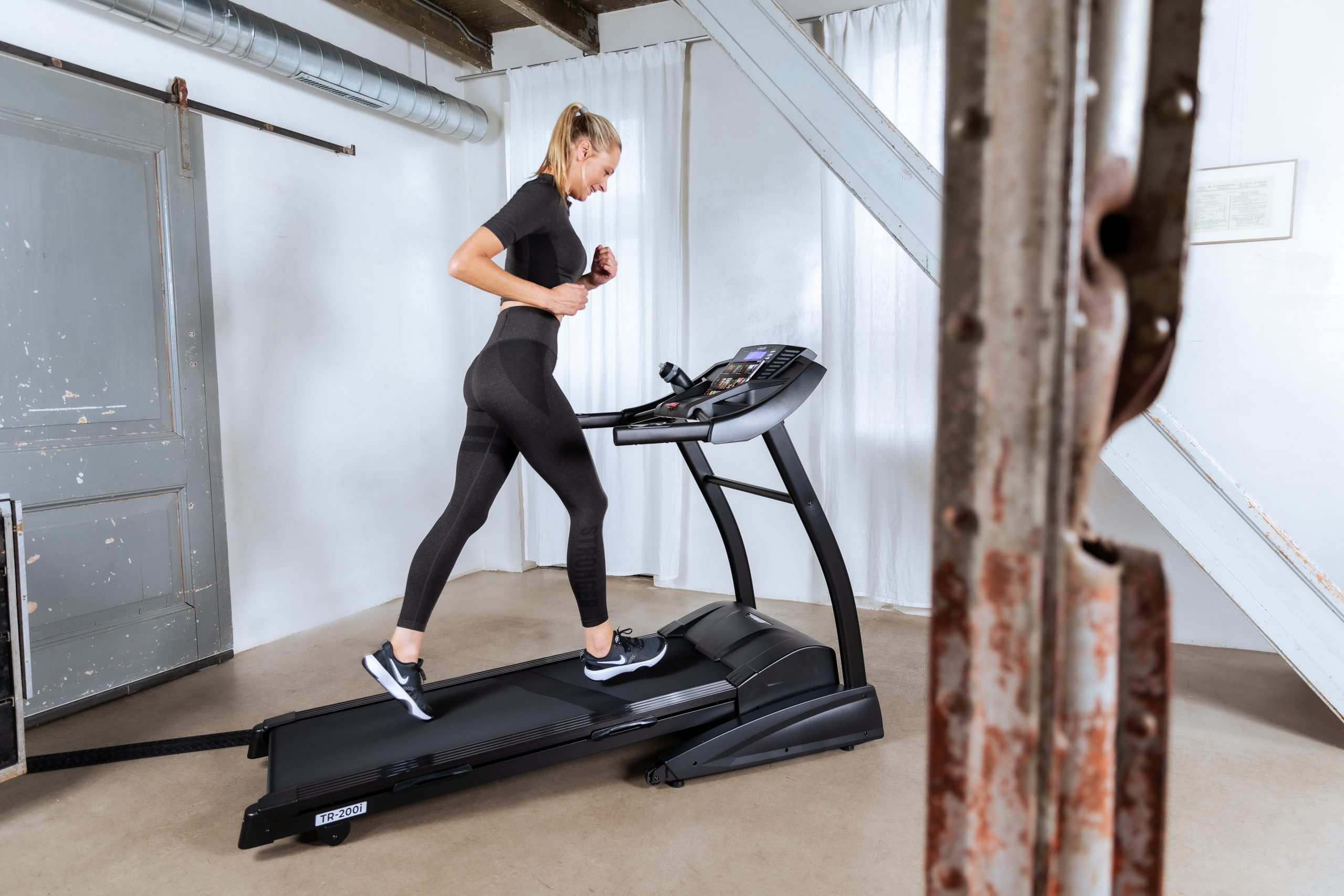 Frau läuft auf einem Laufband in einem modernen Raum. Sie trägt schwarze Sportkleidung.