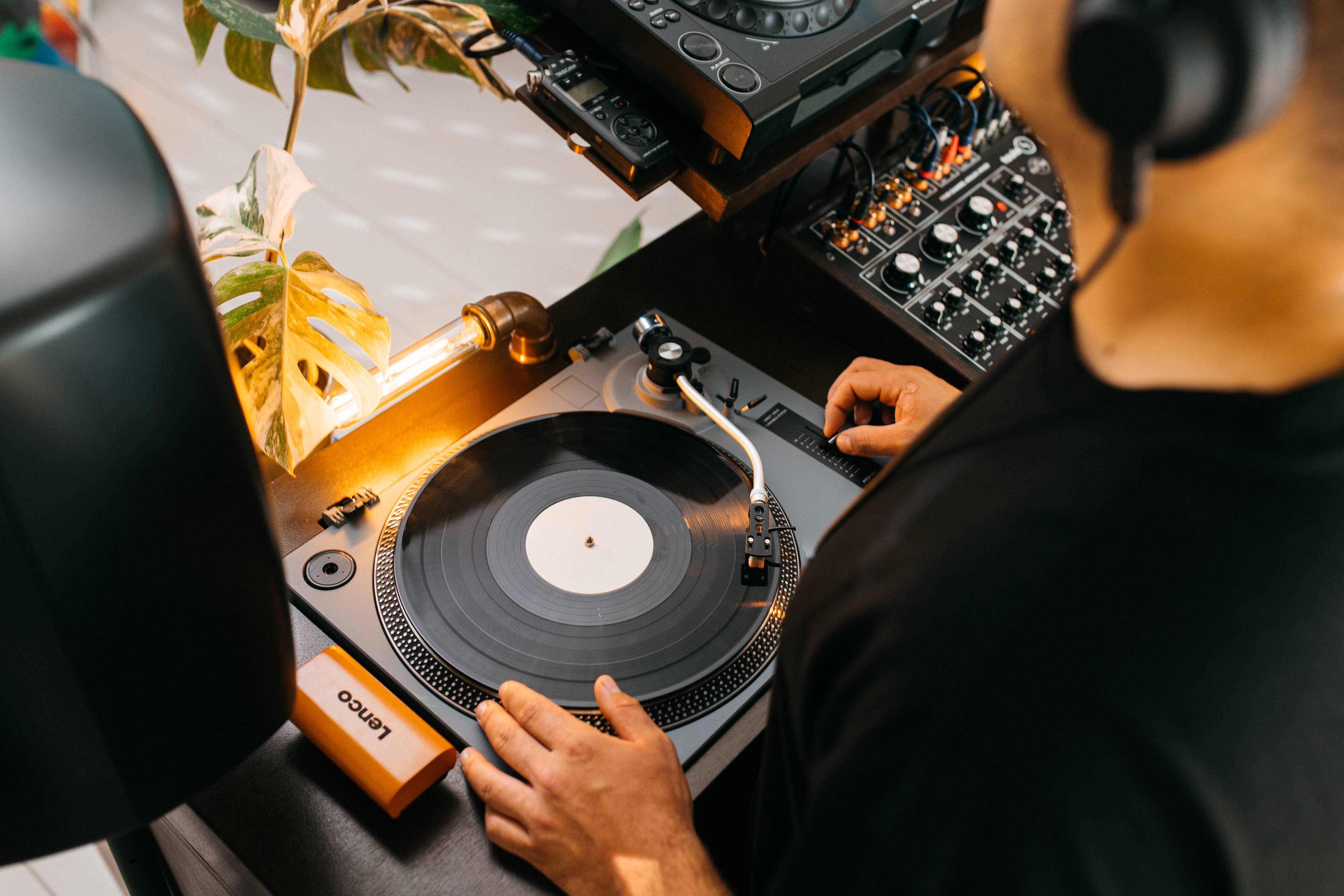 Un DJ usando un tocadiscos. La persona lleva auriculares y utiliza una mesa de mezclas. Una luz y una planta están al lado.