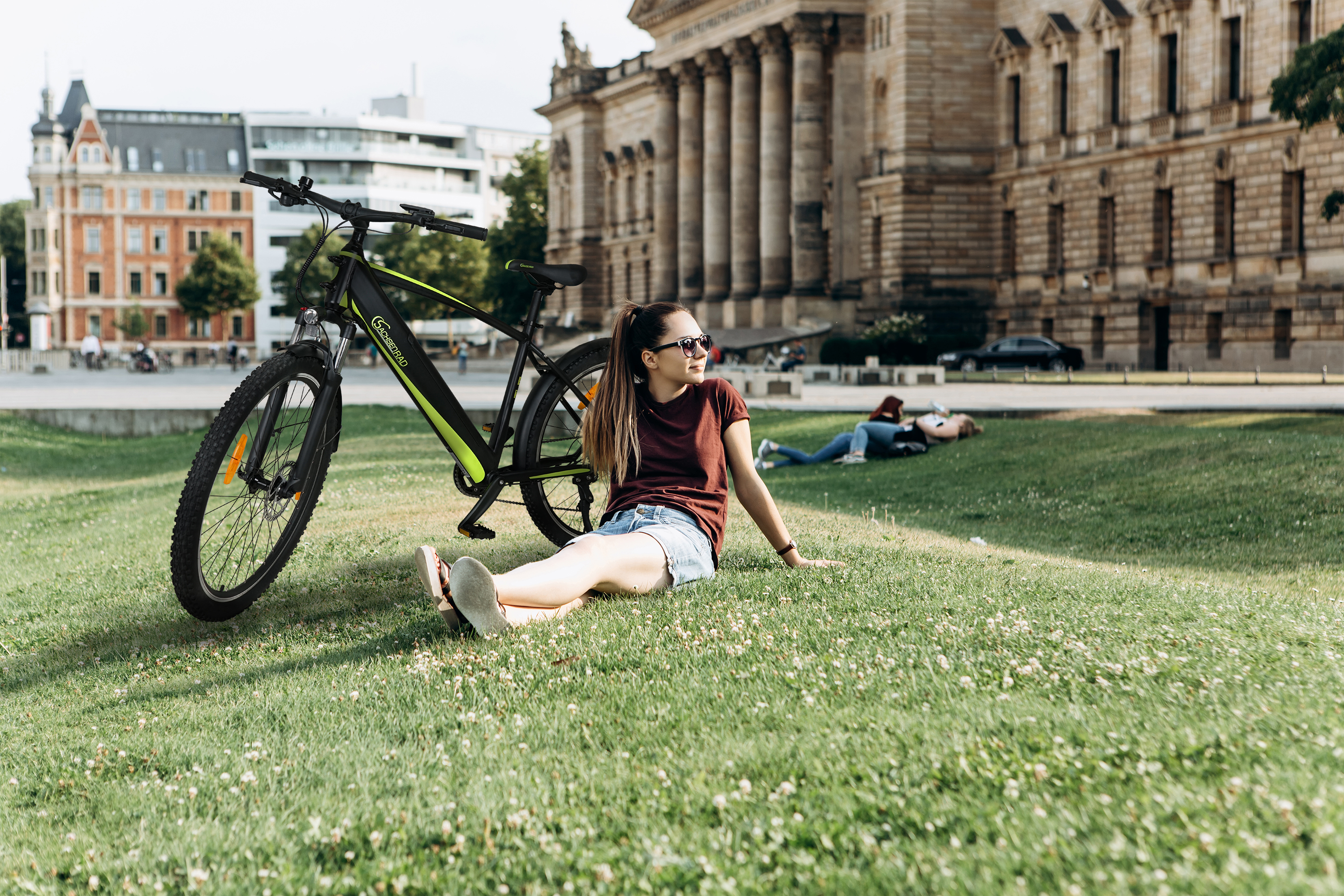 Eine Frau sitzt auf Gras neben einem schwarz-grünen Fahrrad. Zwei weitere Personen liegen auf dem Gras.