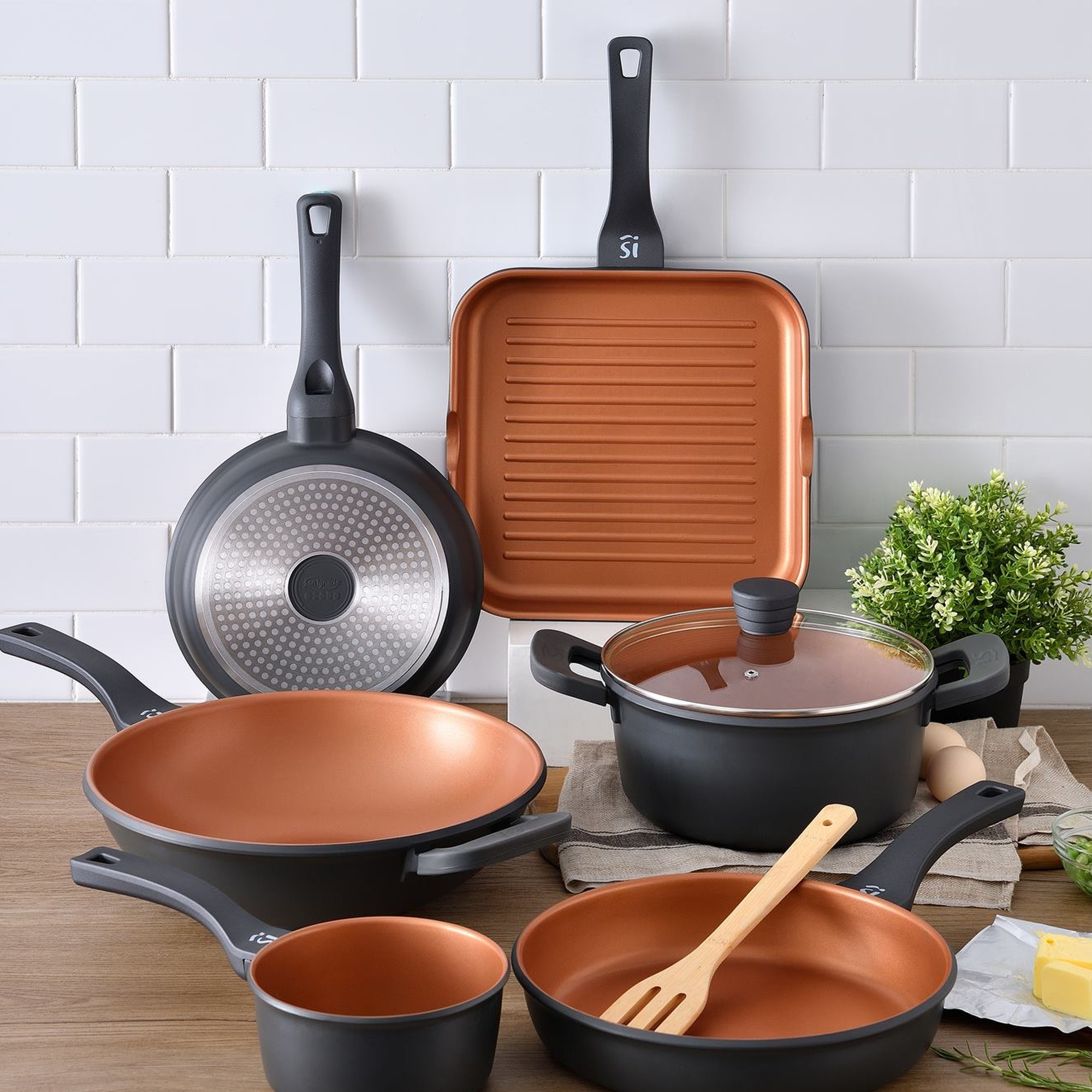 Un conjunto de utensilios de cocina negros y cobrizos sobre una superficie de madera y una pared de azulejos blancos.
