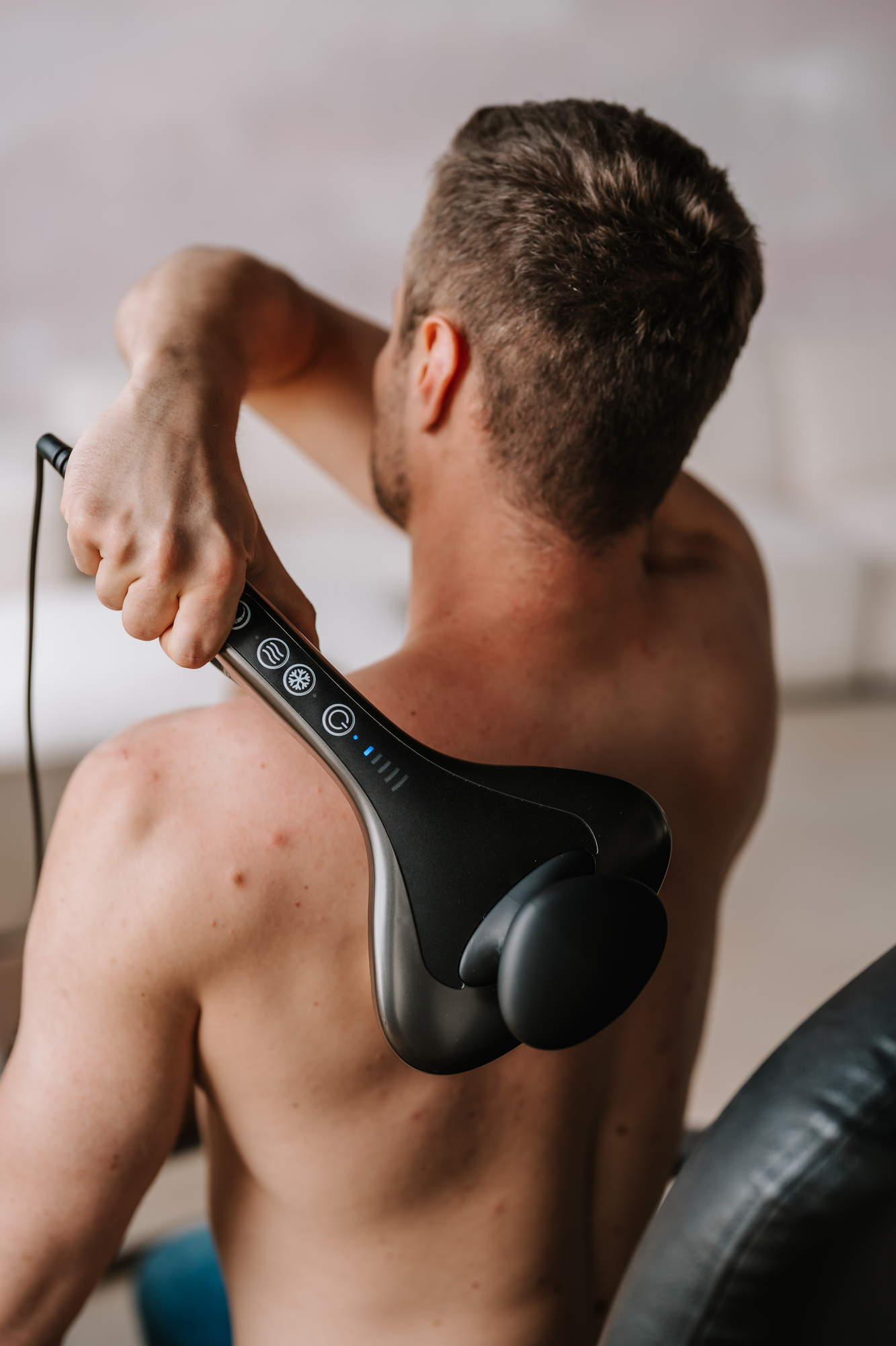 Mann benutzt ein schwarzes Massagegerät am Rücken. Das Gerät ist mit einem Kabel verbunden.