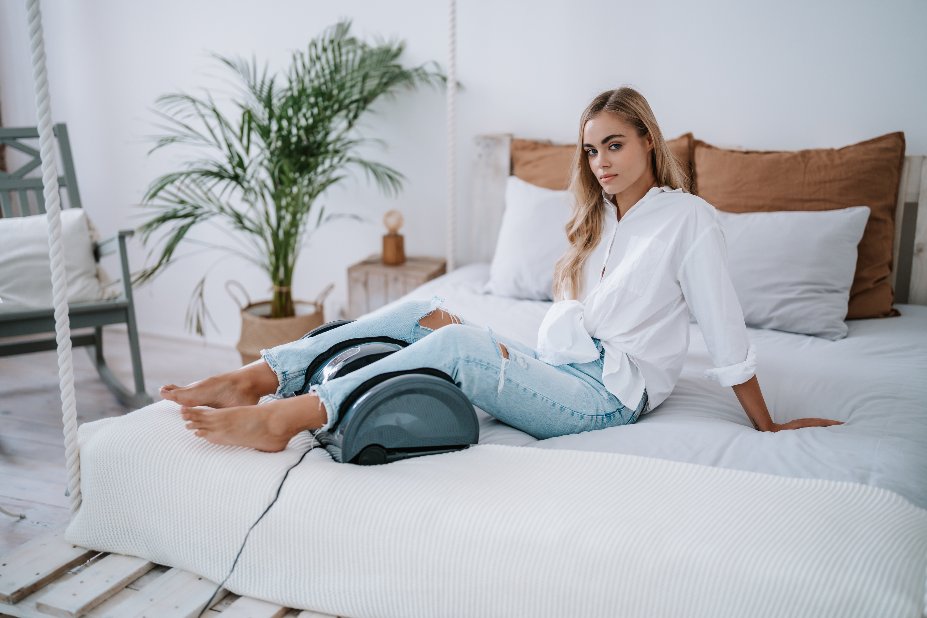 Blonde Frau sitzt auf einem Bett mit einem Fußmassagegerät. Sie trägt ein weißes Hemd und zerrissene Jeans.