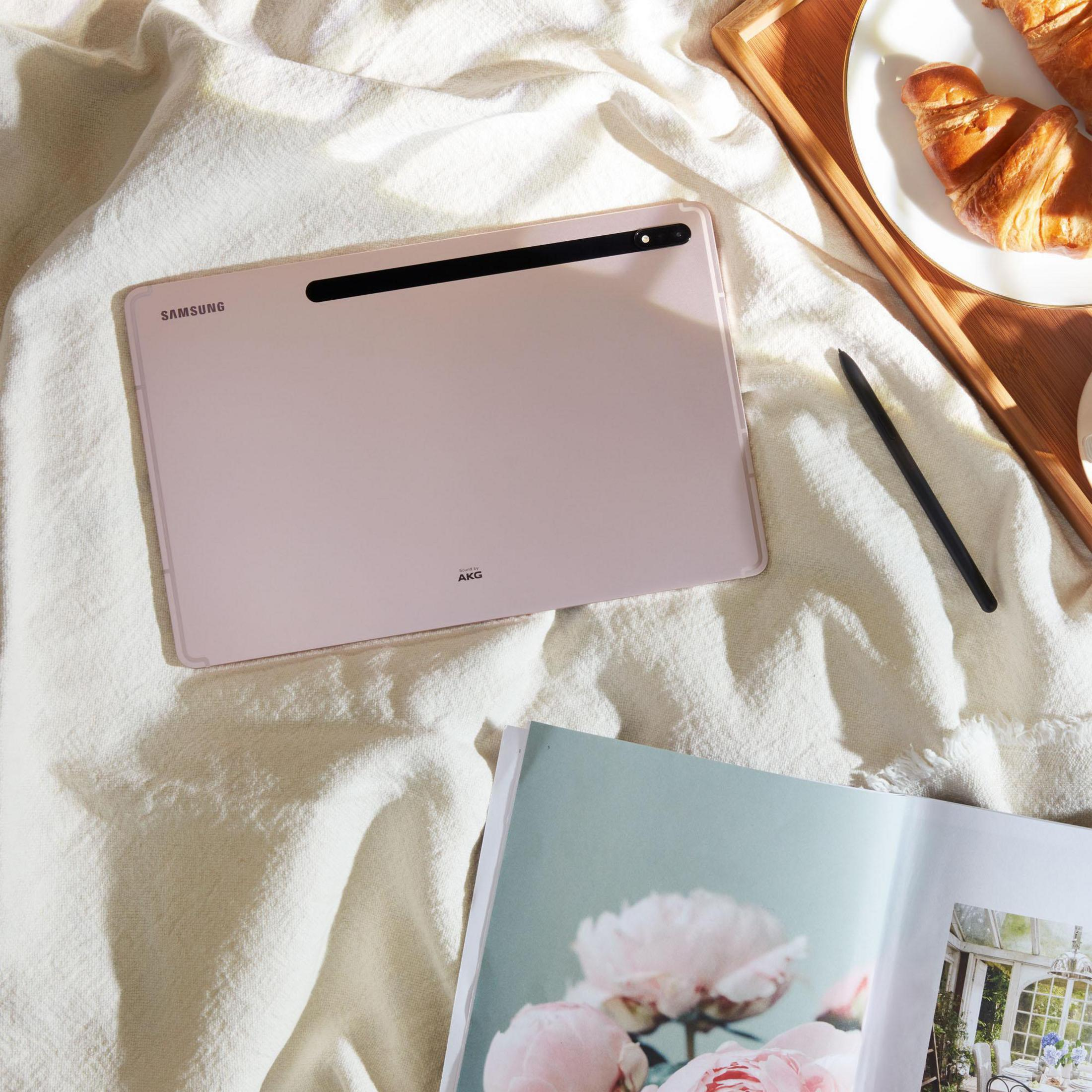Różowy tablet na białym łóżku, z długopisem i talerzem ciast, widok z góry.