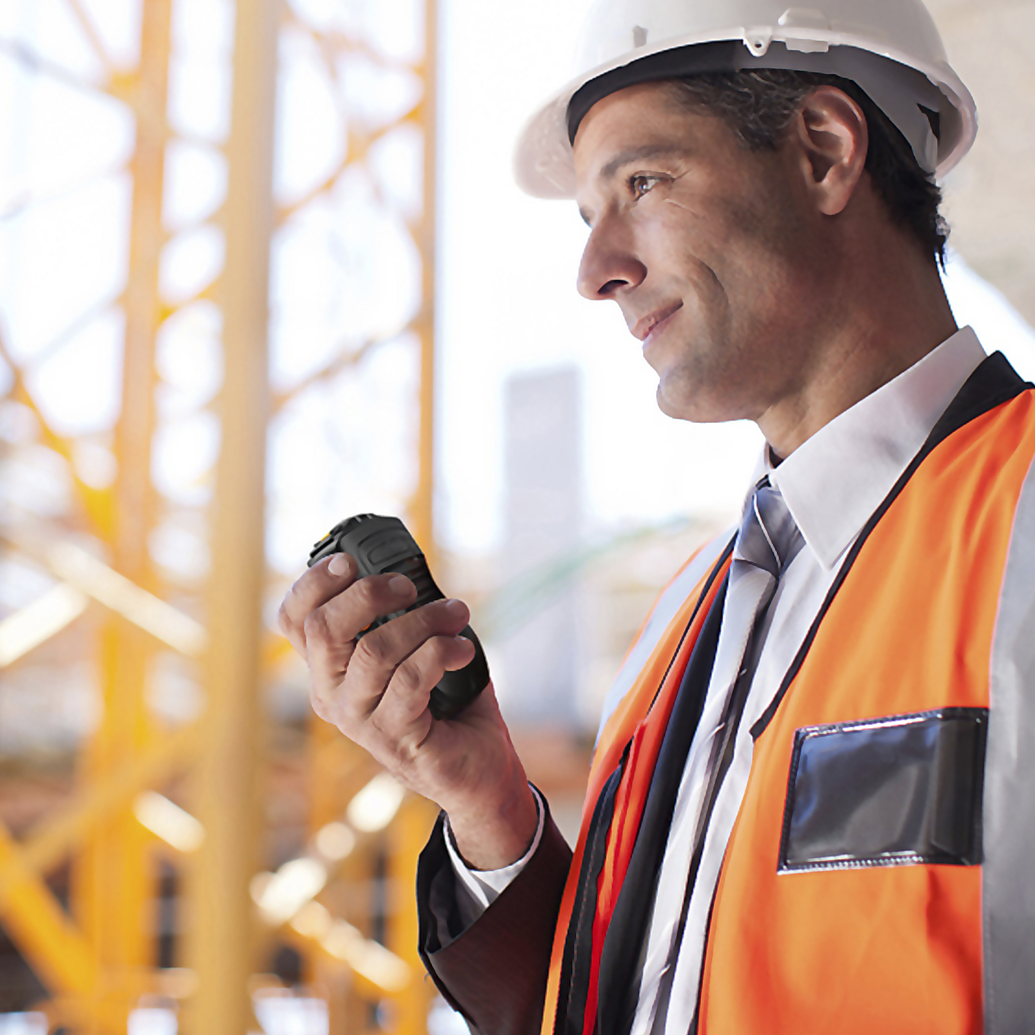 Trabajador de la construcción con chaleco, casco, sosteniendo un walkie-talkie, mirando a la derecha.