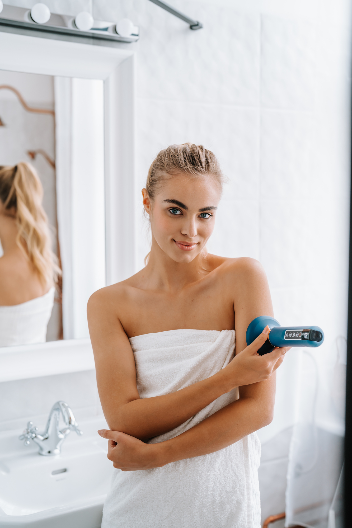 Mujer en toalla blanca sosteniendo un dispositivo de belleza azul. Sonríe en un baño blanco.