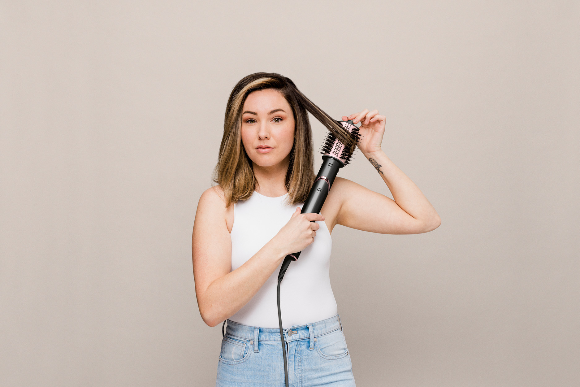 Una donna che tiene in mano una spazzola per capelli, che guarda la fotocamera. Indossa un top bianco e jeans.