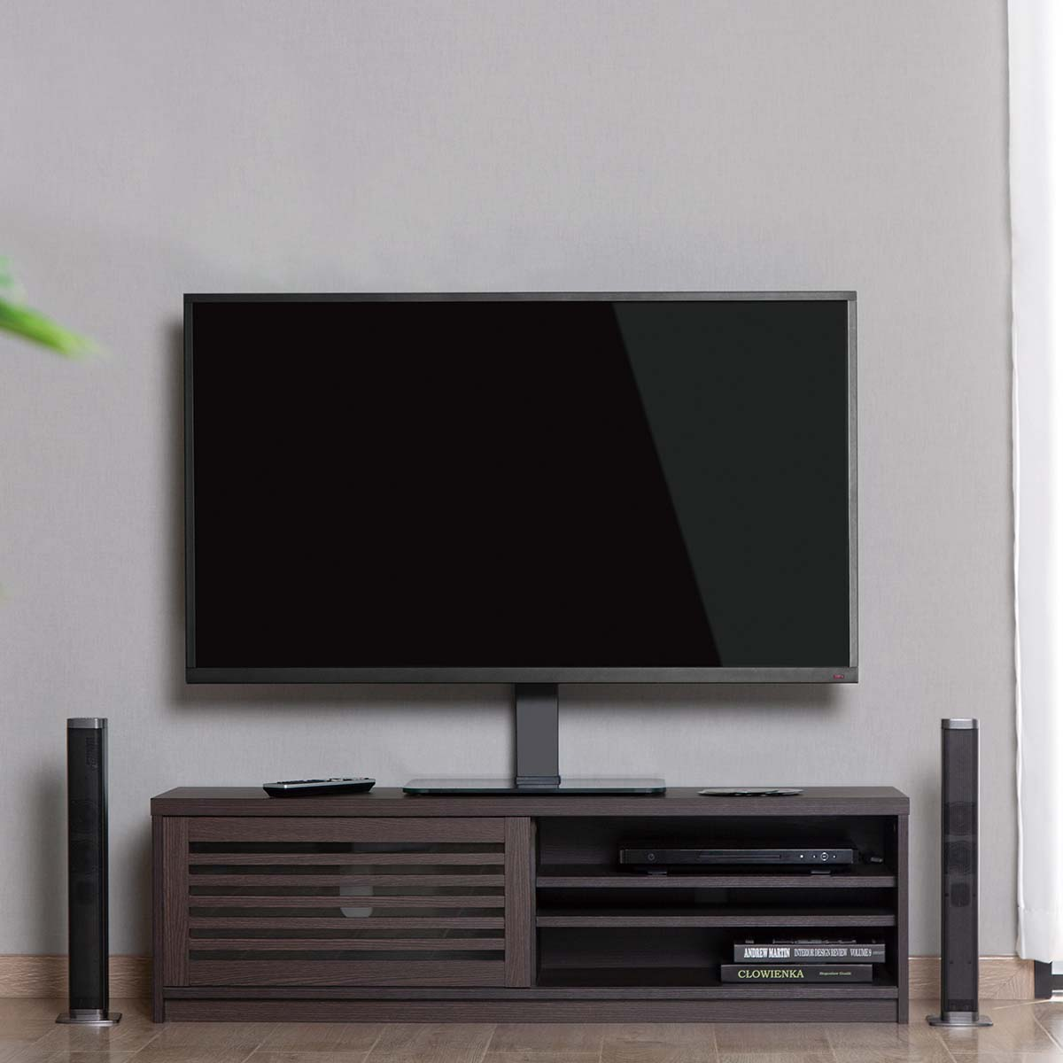 Ein großer schwarzer Fernseher auf einem Ständer, über einem dunklen Holzschrank. Lautsprecher auf beiden Seiten. Grauer Wandhintergrund.
