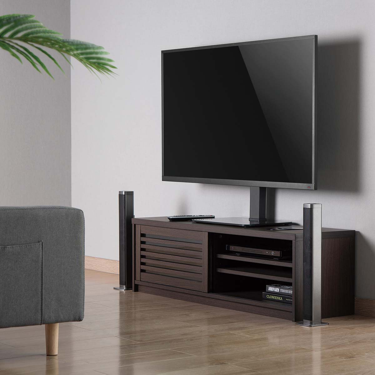 Modernes Wohnzimmer mit großem Fernseher auf Ständer, dunklem Holzschrank und zwei Lautsprechern. Sofa links, Holzboden.