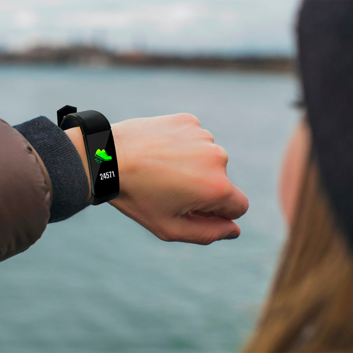 Osoba nosząca czarny smartwatch z zielonymi krokami na nadgarstku. Tło to rozmyta woda i niebo.
