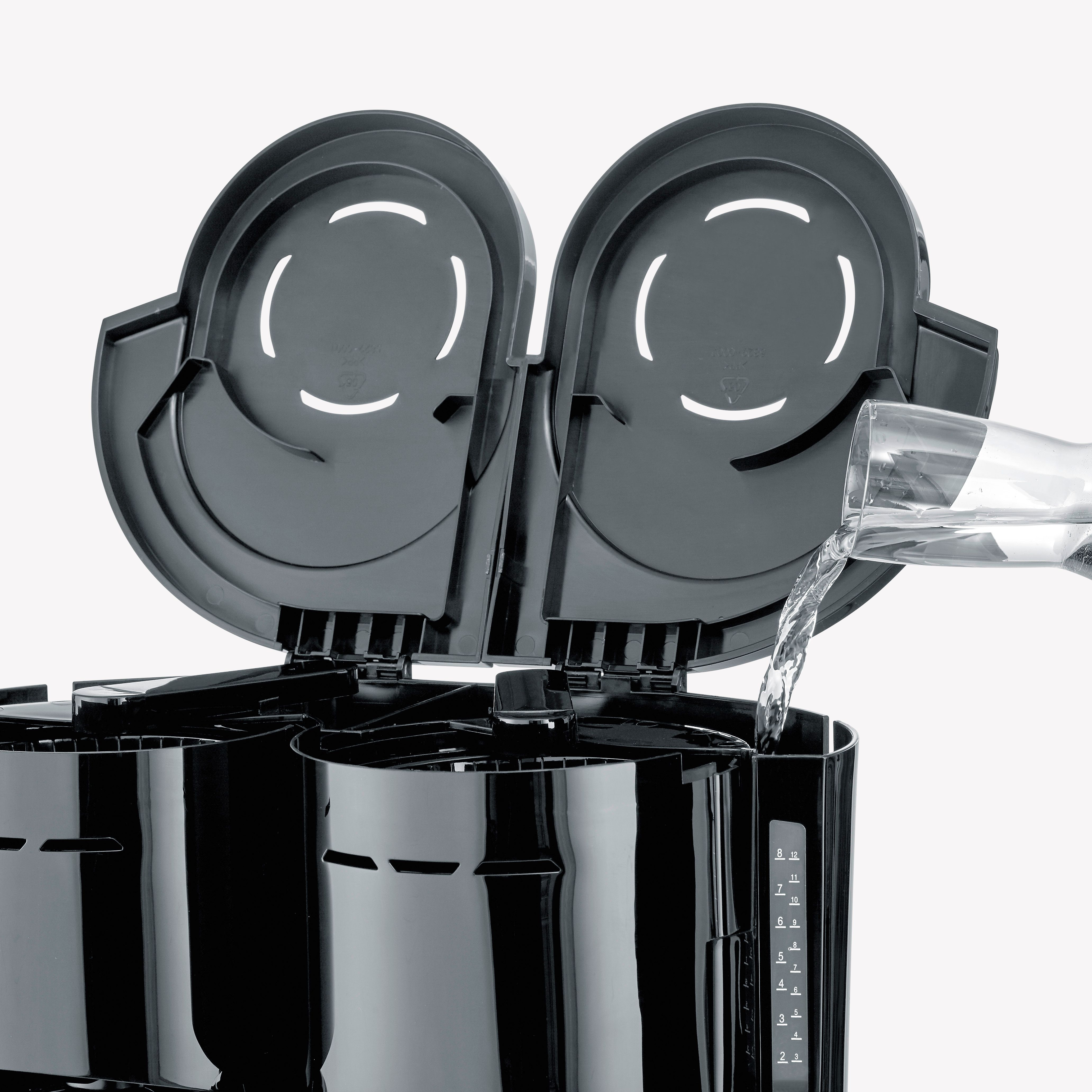 Ekspres do kawy z otwartymi pokrywami, woda wlewana ze szklanego dzbanka do czarnej maszyny.