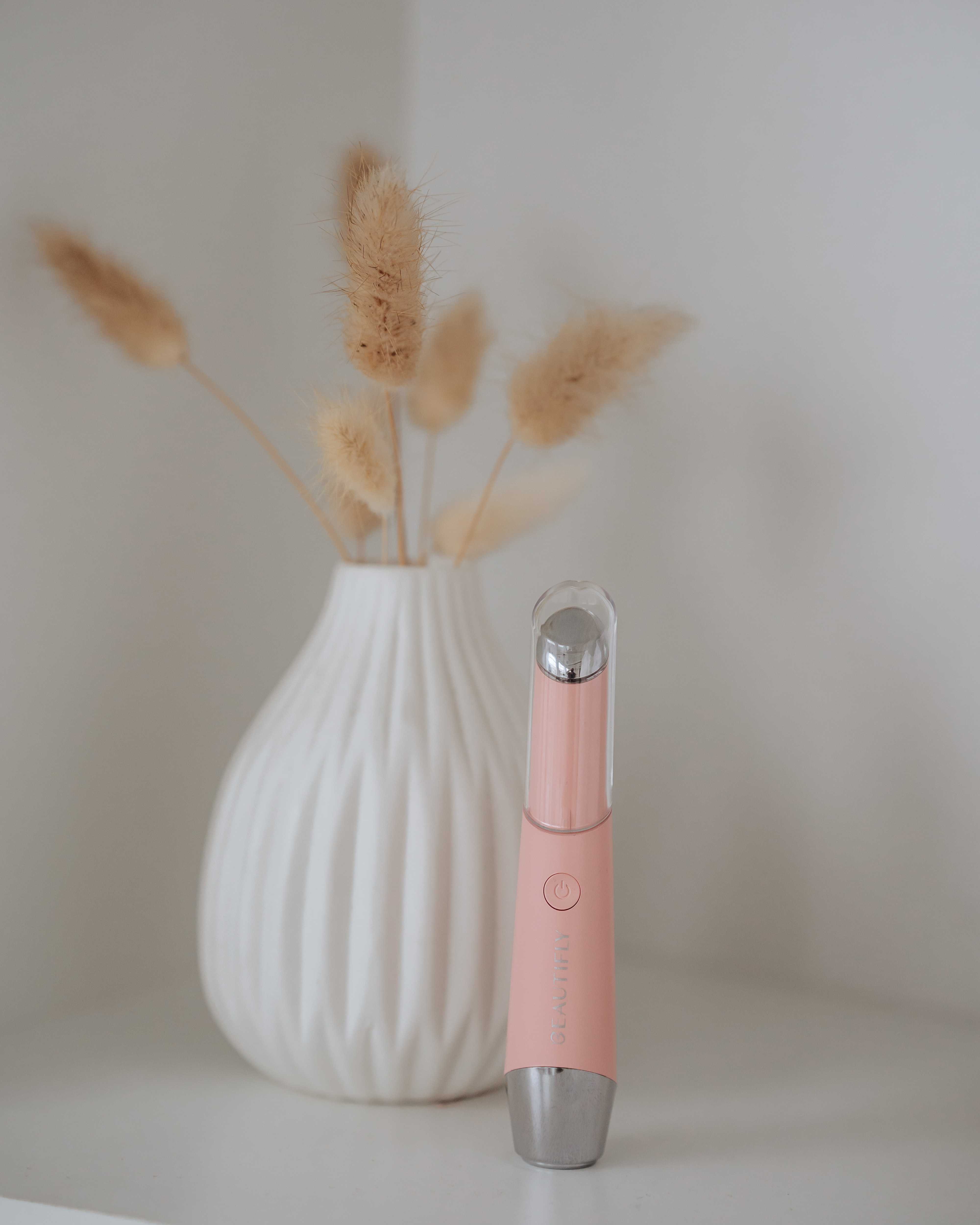 Pinker Augenmassager neben einer Vase mit Trockenblumen auf einem weißen Regal.