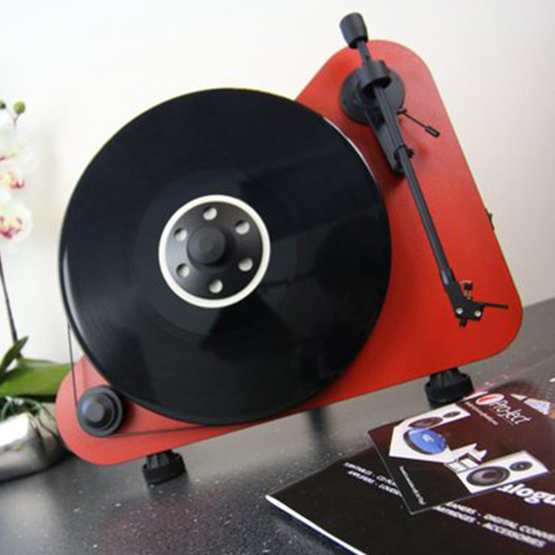 A red turntable with a black record. A record is on the turntable and a few brochures are nearby.