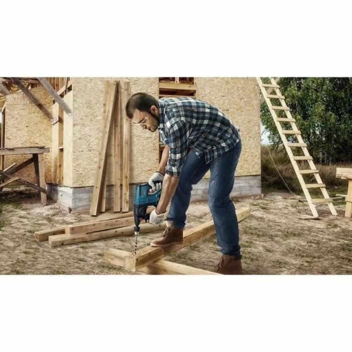 Ein Mann bohrt Holz. Er trägt ein kariertes Hemd und Jeans.