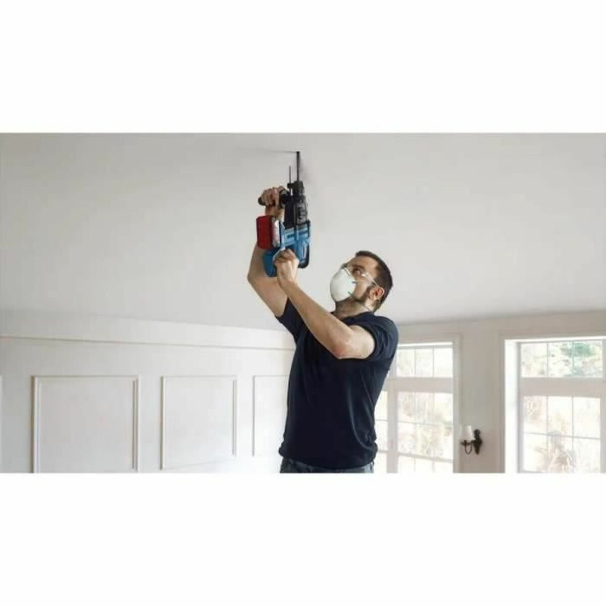 Ein Mann mit Maske bohrt in eine Decke. Er hält einen blau-roten Bohrer.