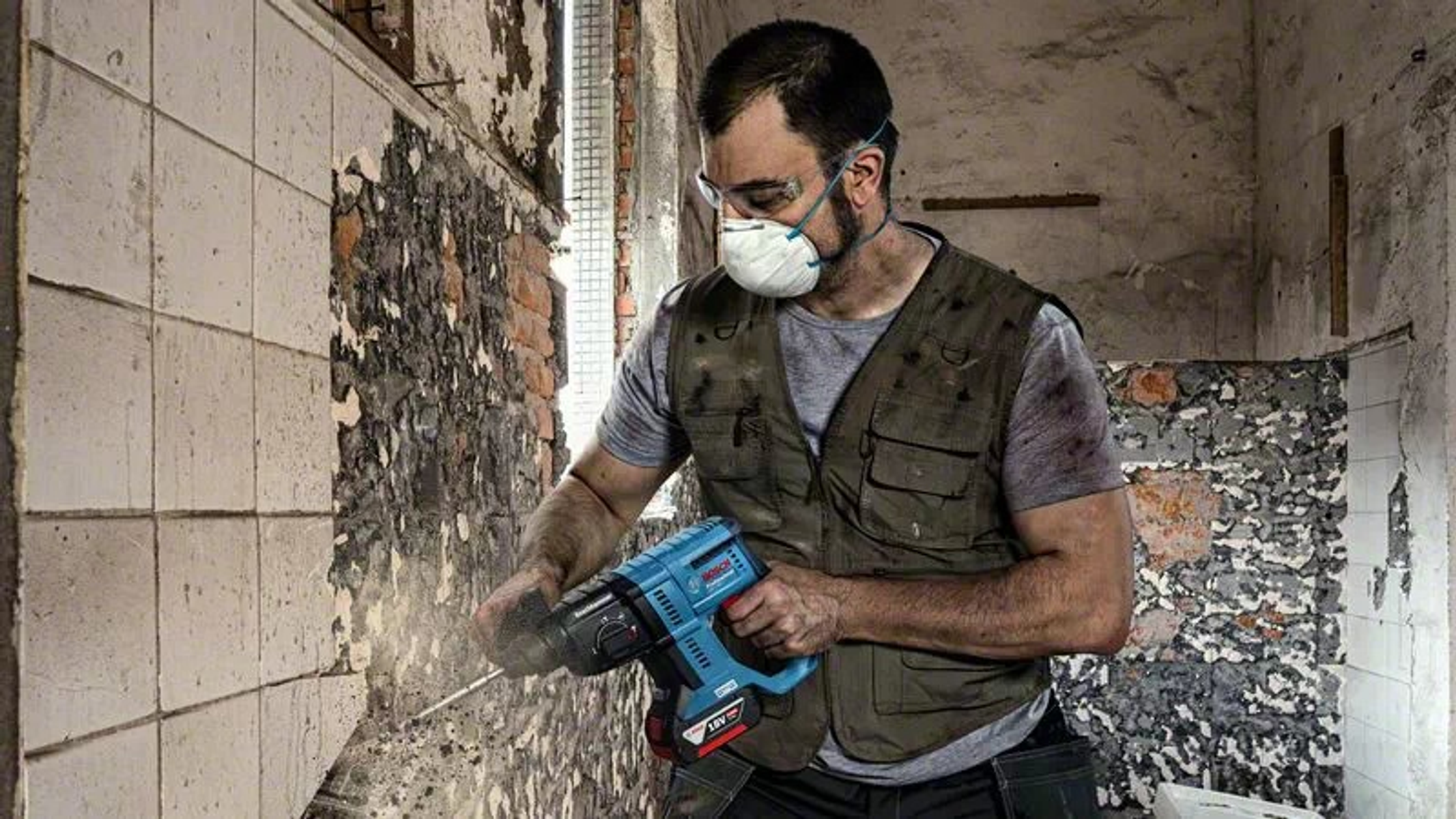 Ein Mann mit Maske und Schutzbrille benutzt einen Bohrer an einer Wand und reißt sie ab.