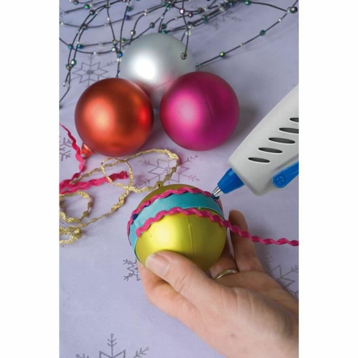 Adornos navideños siendo decorados. Una pistola de pegamento aplica pegamento a un adorno dorado. Mesa morada.