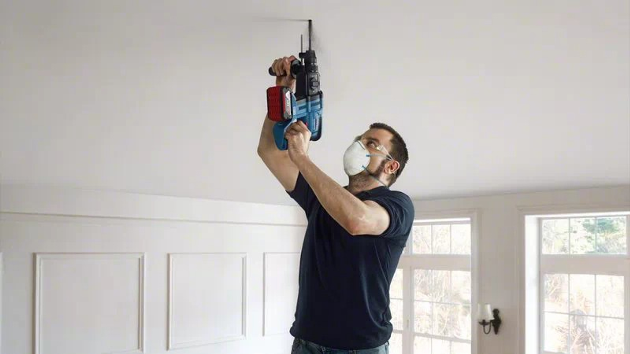 Ein Mann mit Maske bohrt in eine Decke, drinnen. Bohrer und Decke sind weiß.