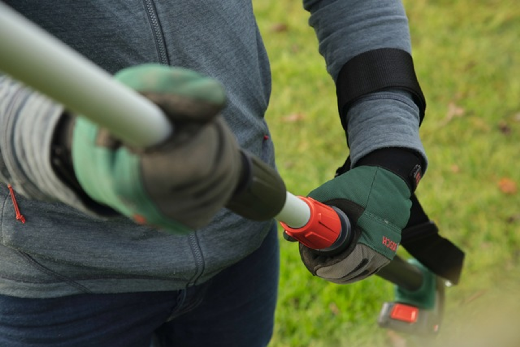 Person hält Gartengerät mit grünen Handschuhen. Werkzeug dunkel mit rotem Knopf. Hintergrund ist Gras.