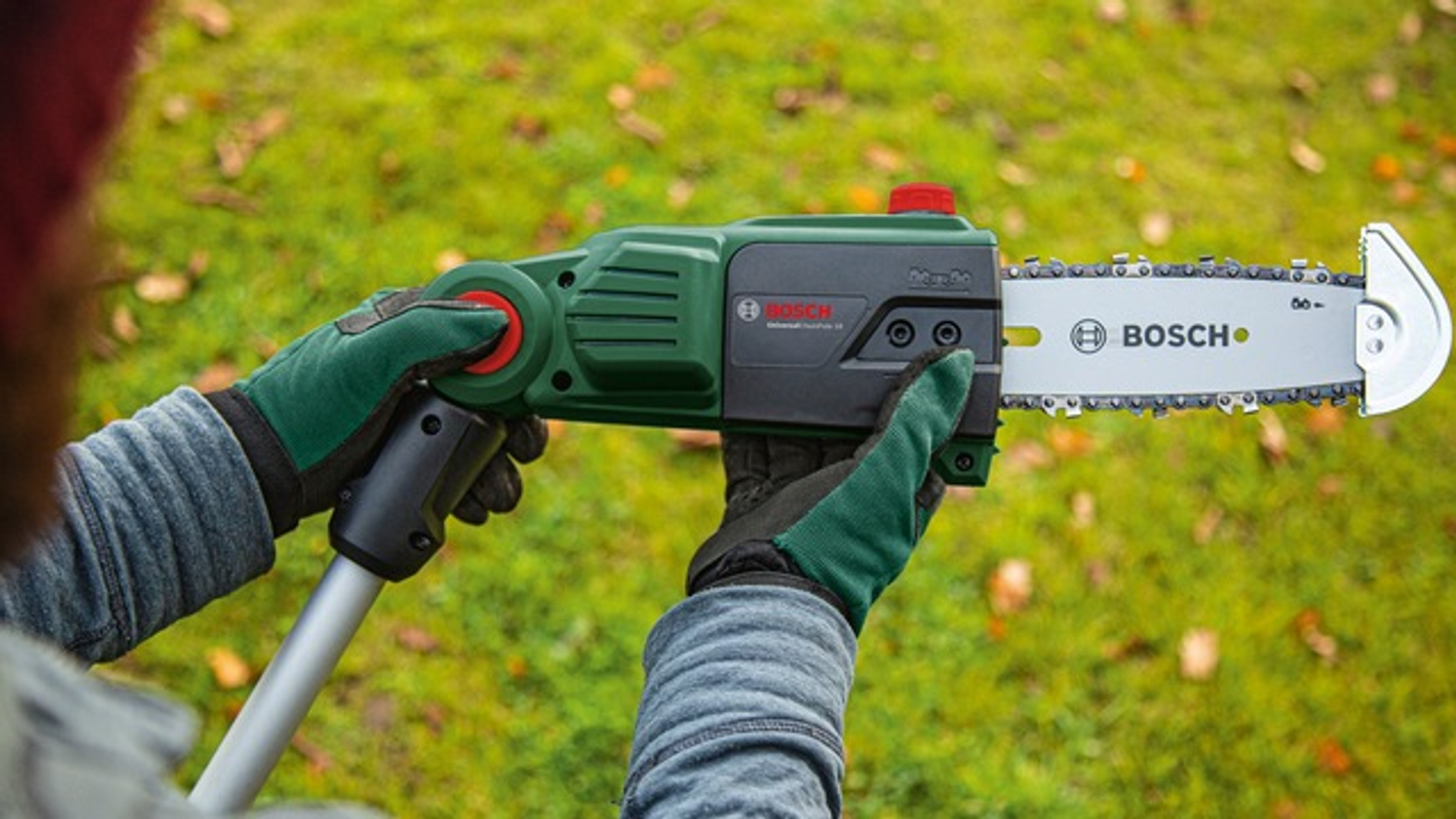 Eine Person hält eine grüne und schwarze Bosch-Kettensäge in einem grasbewachsenen Bereich, mit grünen Handschuhen.