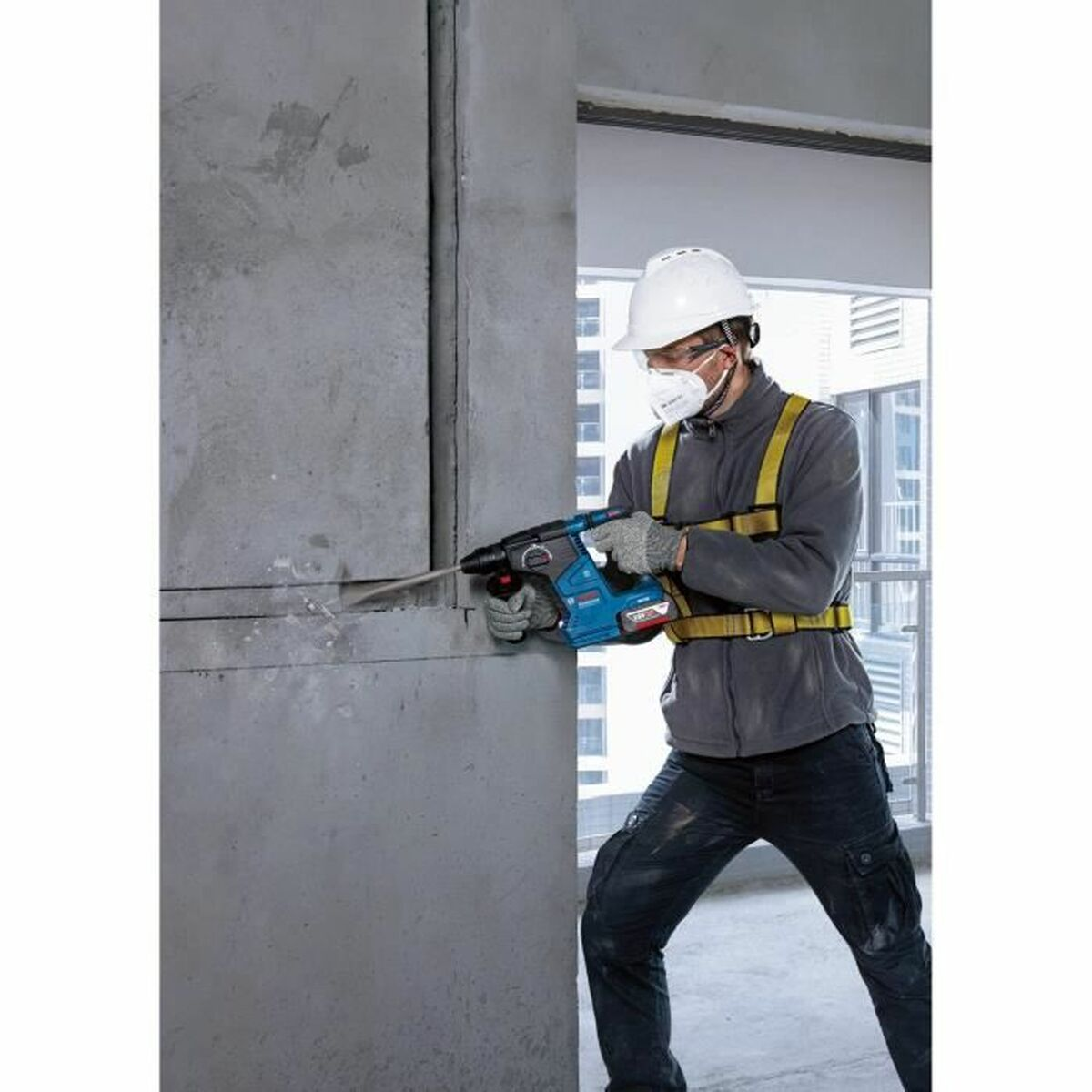 Mann mit Sicherheitsausrüstung, der einen blau-schwarzen Bohrhammer an einer Betonmauer benutzt.