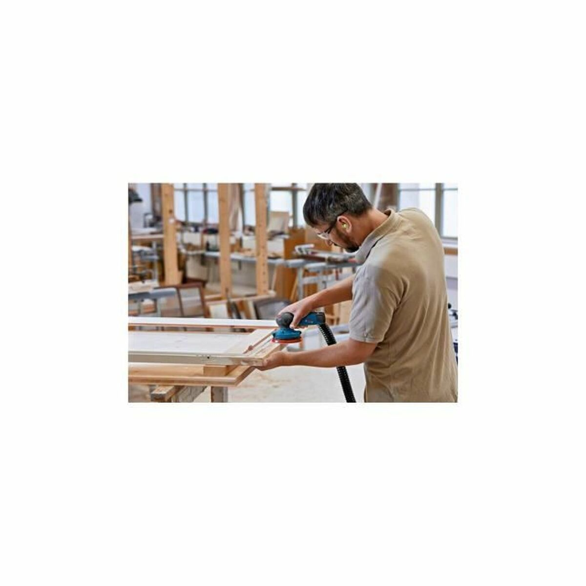 Un homme utilisant une ponceuse sur une planche de bois dans un atelier. Portant des lunettes et une protection auditive.