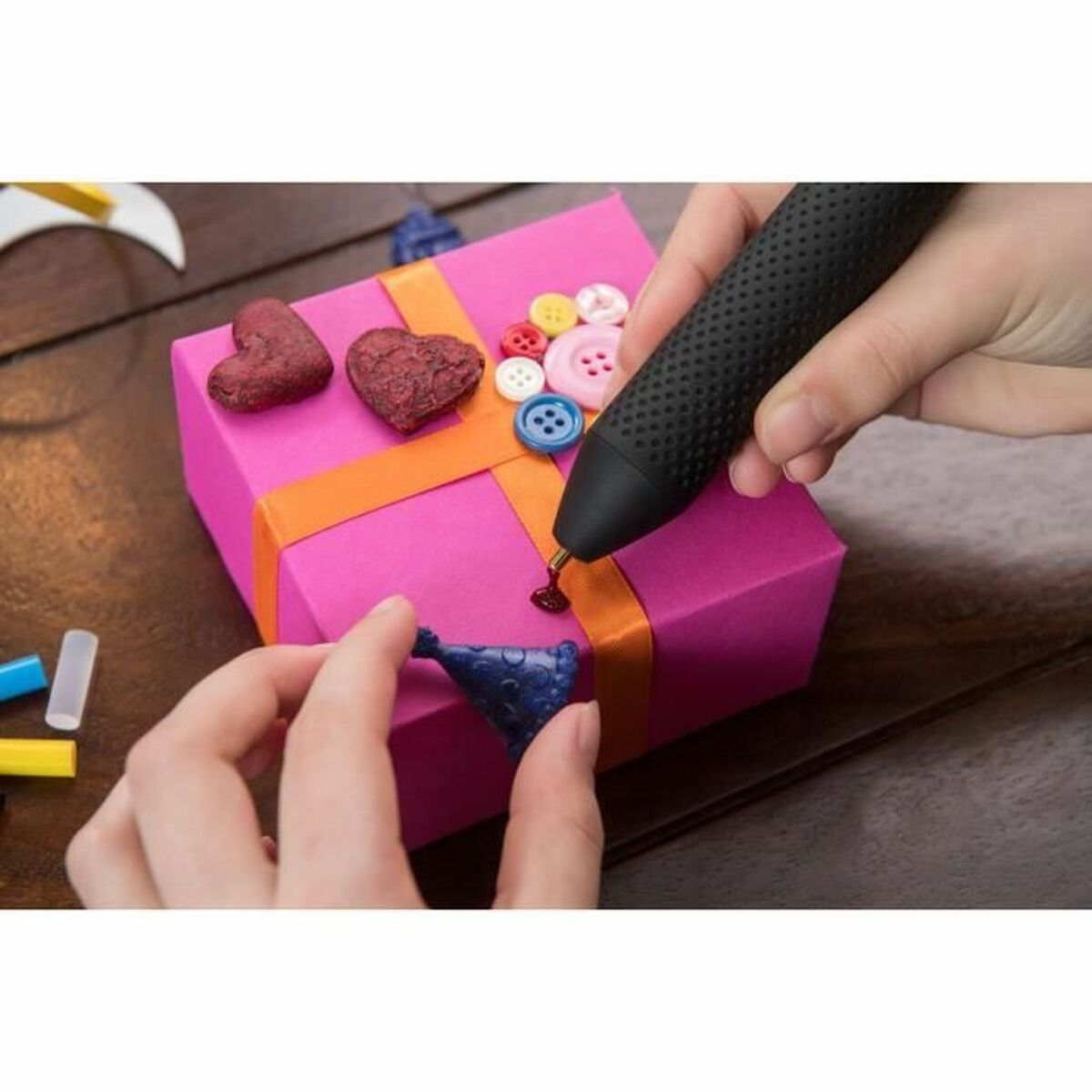 Person verwendet eine Klebepistole auf einer rosa Geschenkbox. Auf der Box befinden sich Knöpfe und Herzen.
