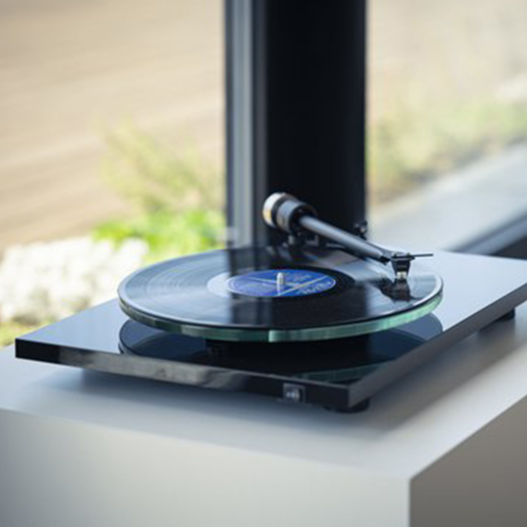 A black turntable playing a record. The turntable is on a white surface near a window.