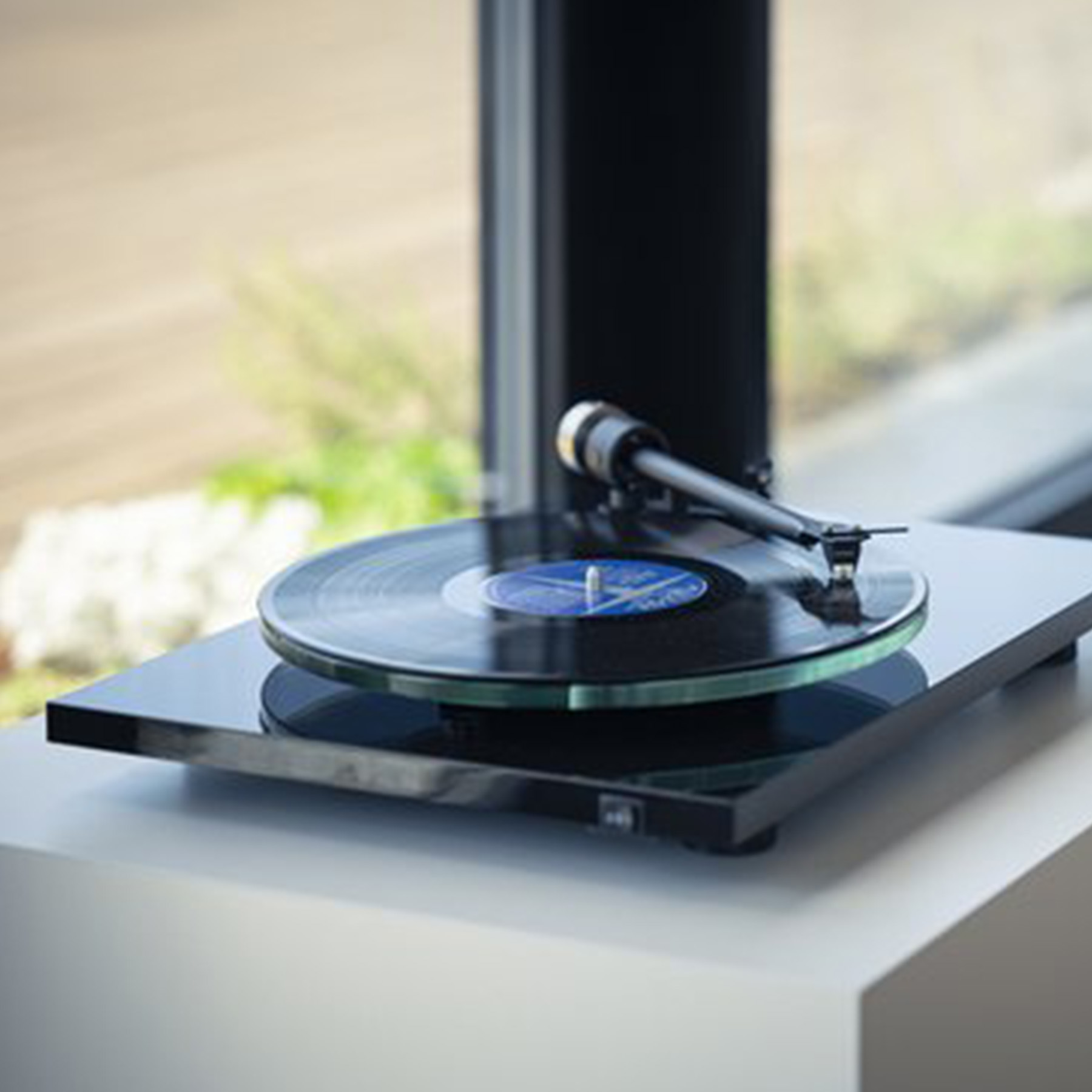 A black turntable playing a record. The turntable is on a white surface near a window.