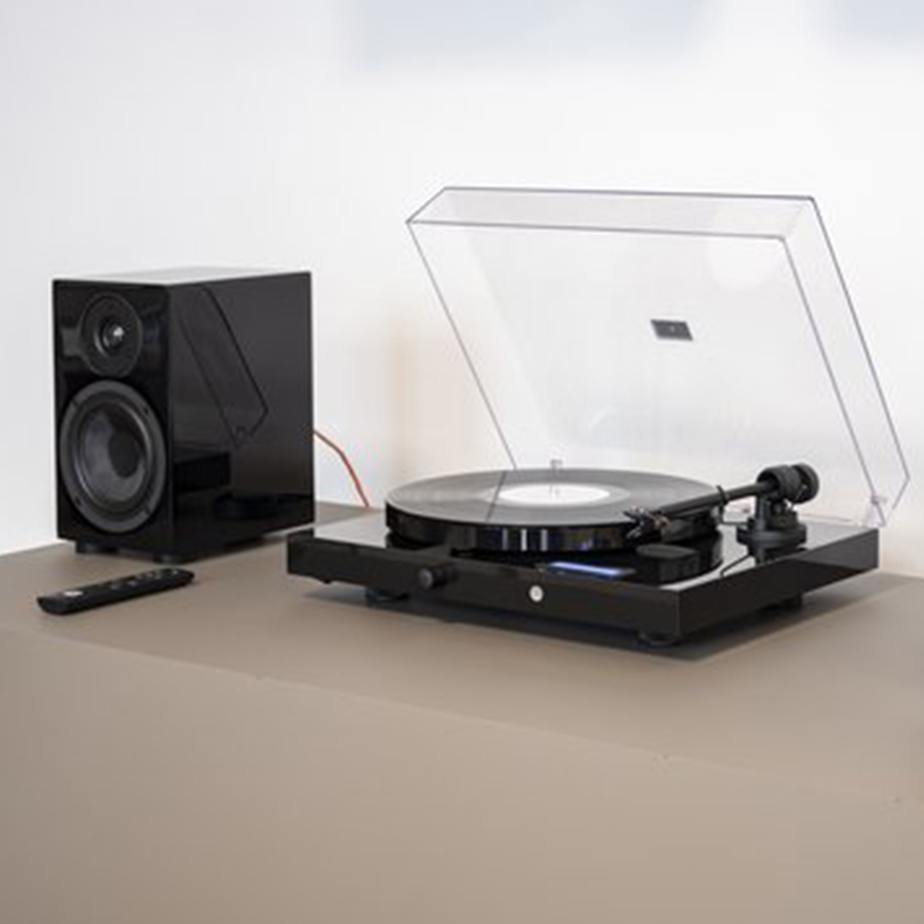 Czarny gramofon, głośnik i pilot na stole. Przezroczysta pokrywa otwarta.