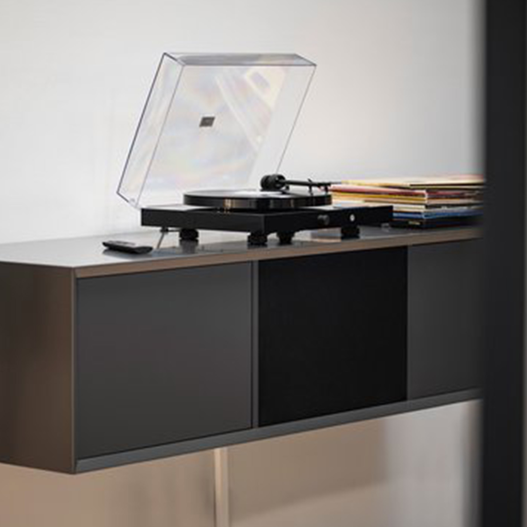 Czarny gramofon z przezroczystą pokrywą, umieszczony na ciemnoszarej szafce. Płyty są ułożone w pobliżu.