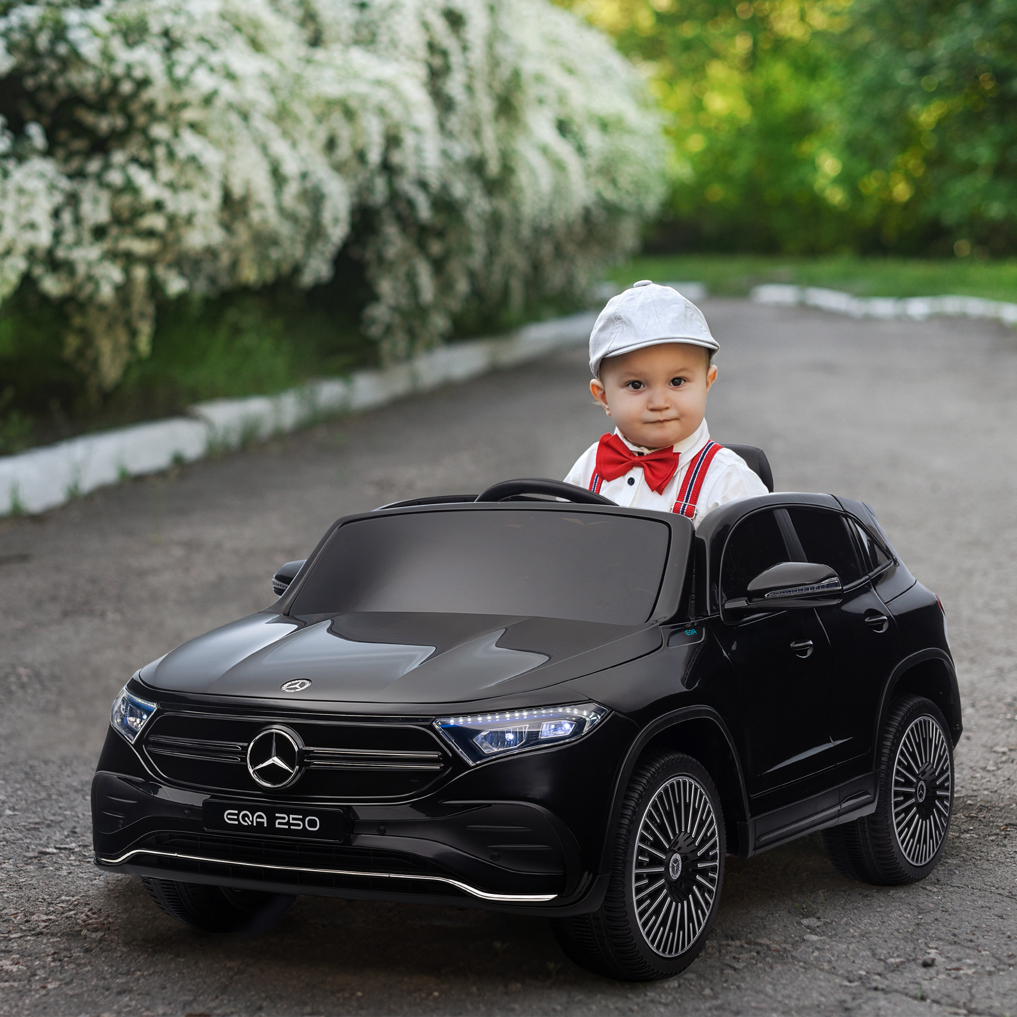 Un bambino in un'auto, con papillon e cappello. L'auto è nera e lo sfondo è una strada e alberi.
