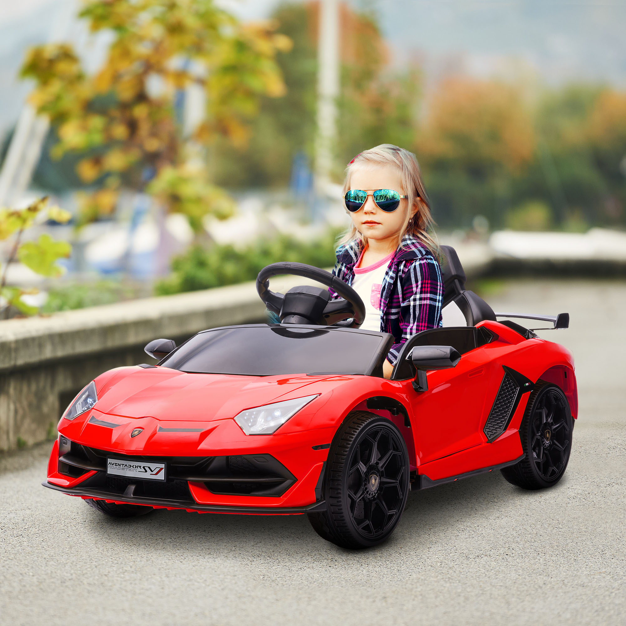 Una bambina con occhiali da sole guida un'auto giocattolo rossa. Strada asfaltata, alberi sullo sfondo.
