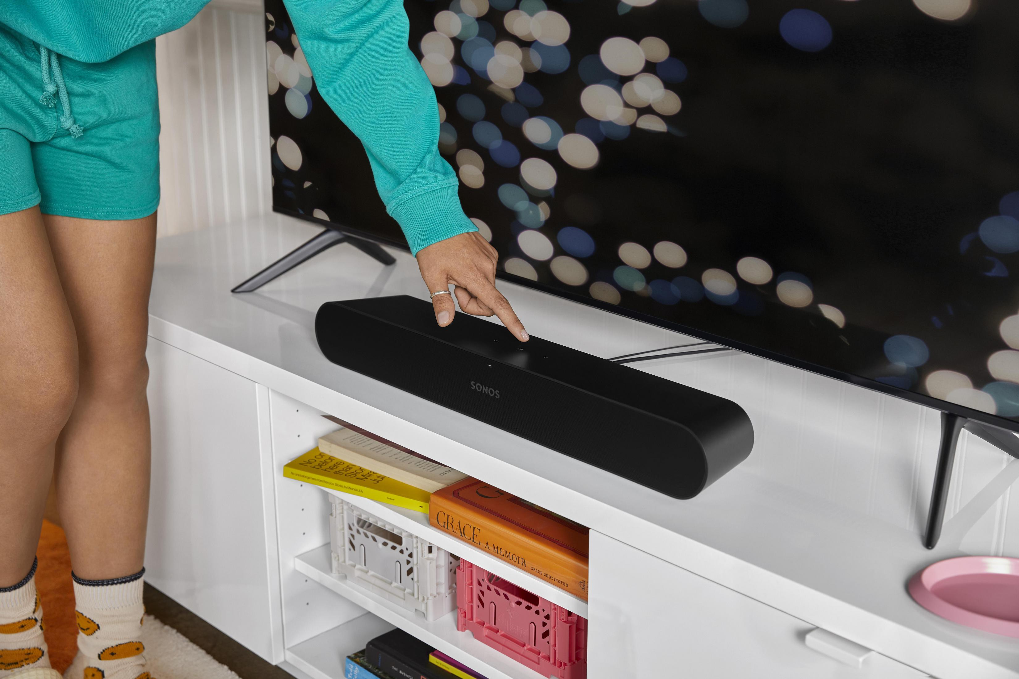 Die Hand einer Person berührt eine schwarze Soundbar auf einem weißen Schrank. Ein Fernseher ist dahinter.