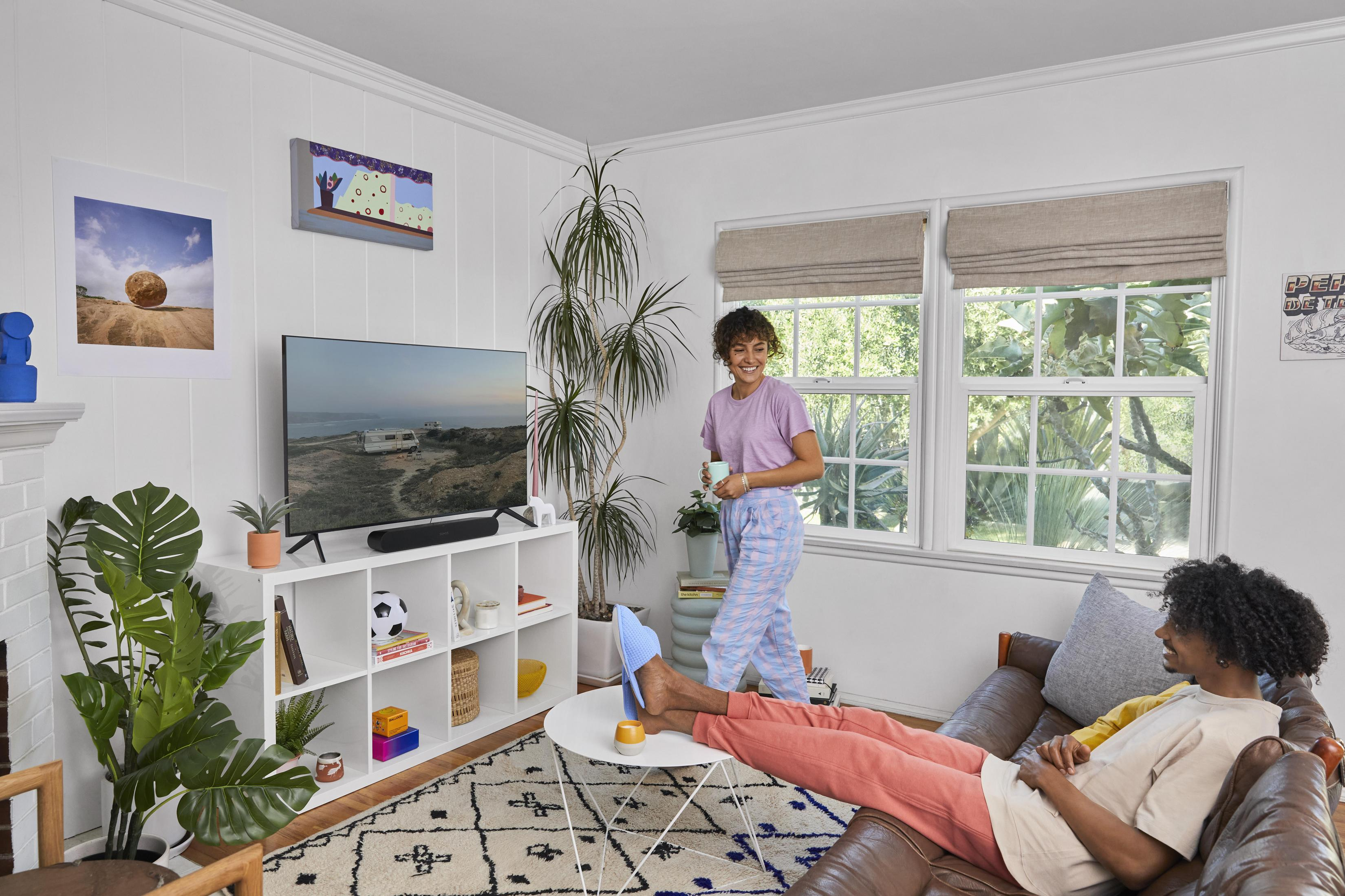 Ein Wohnzimmer mit einem Paar, einem Fernseher, weißen Regalen mit Pflanzen und Kunst an der Wand.